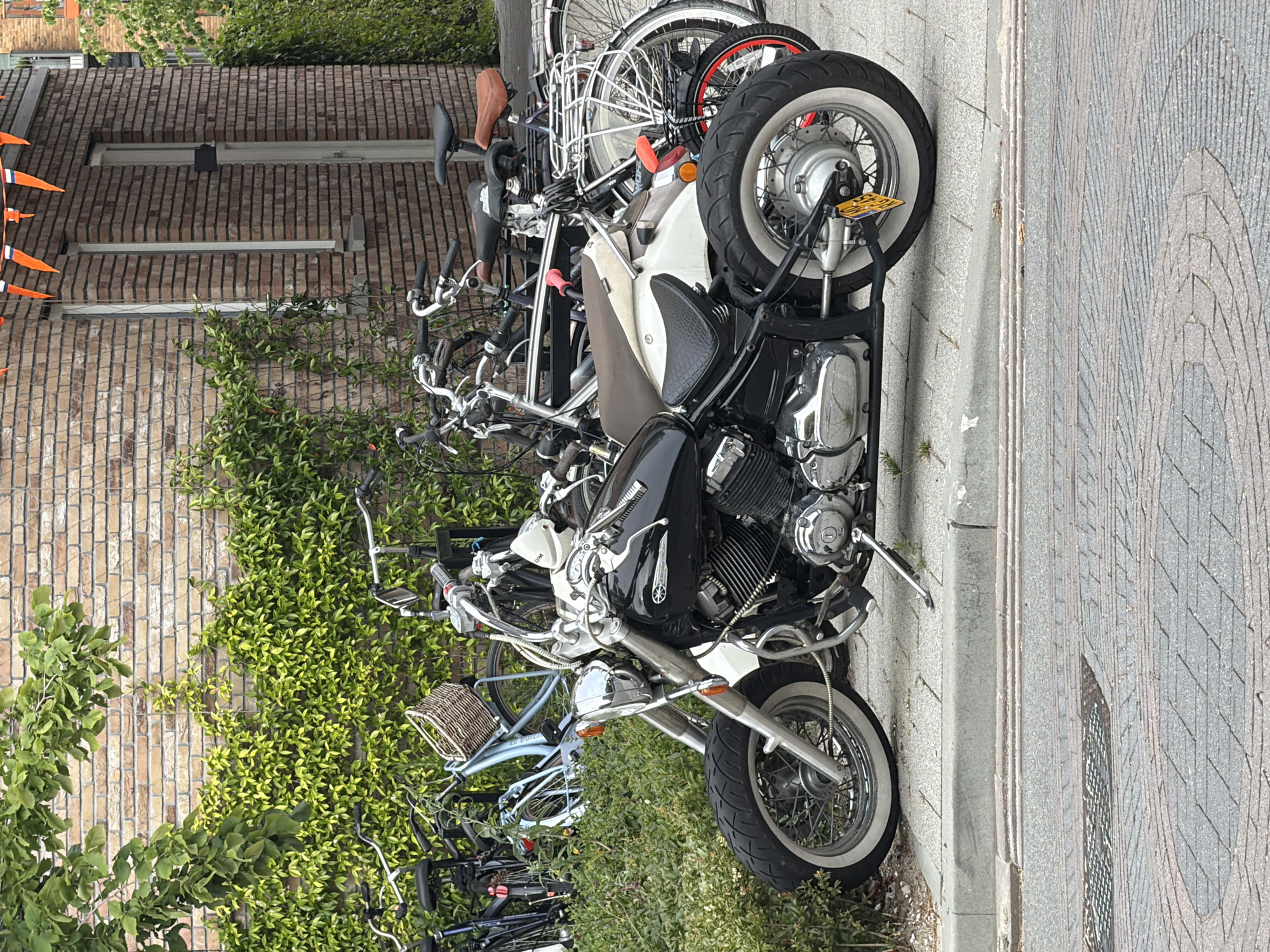 A black motorcycle is parked on a sidewalk in front of a brick building. The motorcycle is a custom cruiser style with chrome accents and whitewall tires. Behind the motorcycle, several bicycles are parked. The building is made of red brick and has ivy growing on it. There are orange and white triangular flags hanging from the building. The sidewalk is made of grey concrete, and the street in front of it is paved with brick. The scene appears to be in a residential area of Rijswijk, Netherlands.