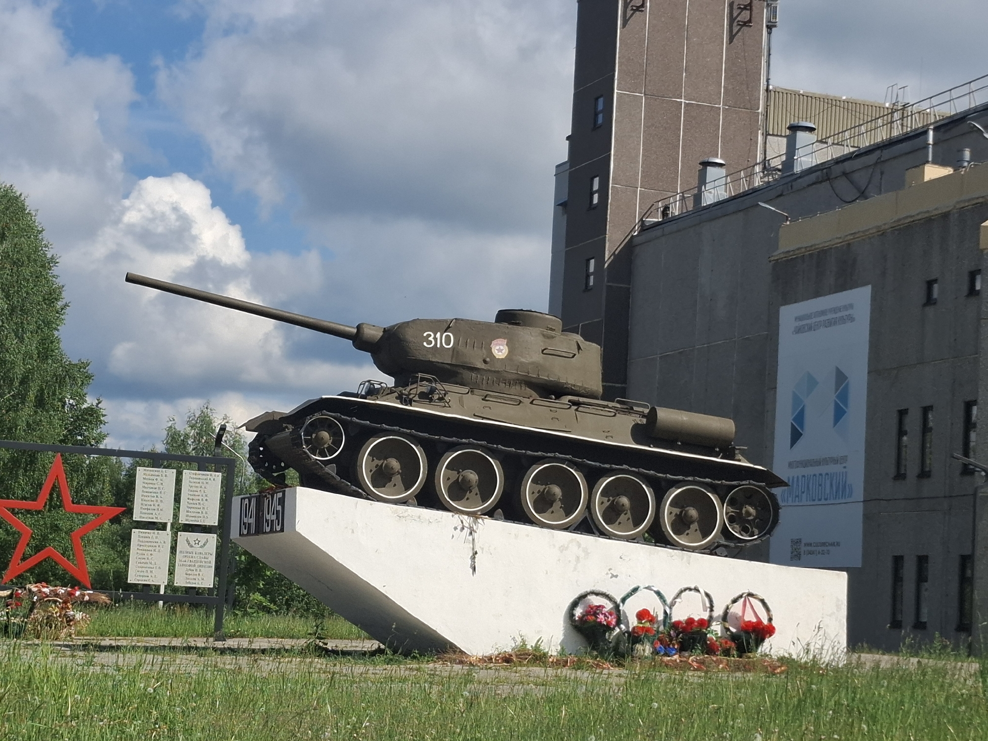 A daytime shot captures a T-34-85 tank, mounted as a war memorial, under a partly cloudy sky. The dark green tank, prominently displaying the number "310" and a red Soviet star insignia on its turret, rests on an angled white concrete pedestal. The years "1941 1945", signifying the Great Patriotic War, are inscribed on the pedestal.
In the foreground, green grass covers the ground, with several wreaths featuring red flowers placed at the base of the tank's monument. To the left, a large red star sculpture stands alongside multiple white plaques listing names in Cyrillic, presumably of fallen soldiers, with one plaque bearing the inscription "ПАМЯТНИК ВОИНАМ..." (Monument to soldiers...). Lush green trees are visible to the left and behind the memorial plaques.
The background features a large, multi-story industrial-style building with brown and gray panels and numerous small windows. A prominent billboard on the building displays blue geometric shapes and Cyrillic text, including "МАРКОВСКИЙ", "КАДРЫ. РАБОТА. БУДУЩЕЕ." (Personnel. Work. Future.), and a website address "www.судомеханик.рф", along with a QR code. The bright conditions and the nature of the memorial suggest a solemn atmosphere of remembrance. The overall scene indicates a historical monument in a town or industrial area, consistent with the given location context of Марковский, Russia.