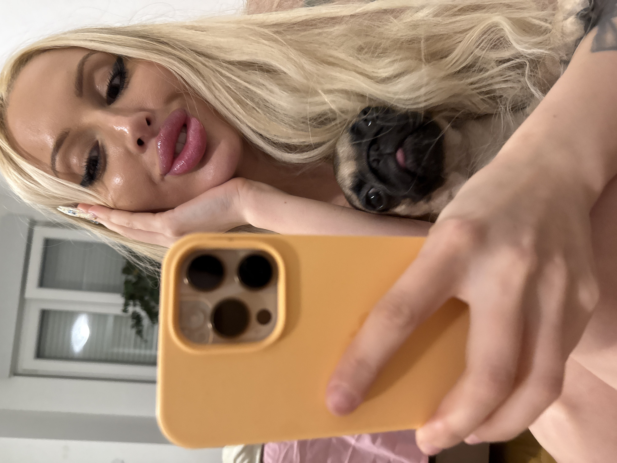 A fair-skinned blonde woman with heavy makeup, including large, glossy lips and long lashes, is holding up a mustard-yellow smartphone to take a selfie with a pug. The pug is partially visible, nestled close to the woman. The woman is holding her head with her left hand. Her left arm has a dark tattoo on it. The background shows a window and some greenery. The location is unknown.