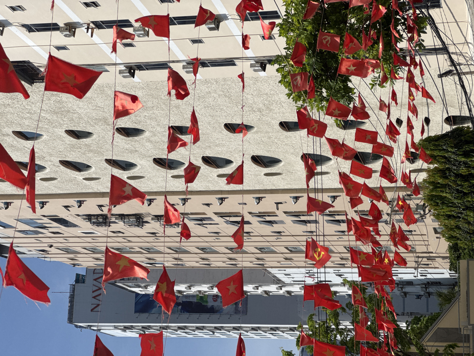 The image depicts a street scene in Bac Nha Trang Ward, Vietnam, adorned with numerous red flags featuring yellow stars and hammer and sickle emblems. These flags are strung across wires, creating a festive or commemorative display. The background consists of modern buildings, one of which is a tall, light-colored structure with a sign that appears to read "NAVADA." Another building is a darker, taller structure with a billboard visible on its side. The flags are the primary focus, with many visible in the foreground and midground, fluttering in the breeze. Trees with green foliage are also present at the bottom of the image, partially obscuring the street level. The sky is a clear blue. There are no people visible in the image.