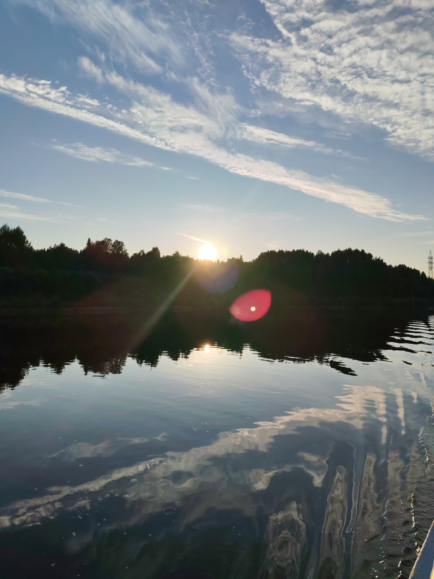 A tranquil river scene unfolds under a bright, sun-drenched sky. The sun, positioned low on the horizon, casts a warm glow across the water, creating a shimmering reflection. A line of trees, silhouetted against the sky, forms a dark band along the riverbank, punctuated by a solitary electricity pylon in the distance on the right. The water's surface is alive with the gentle ripple of waves, reflecting the wispy white clouds that drift across the vast blue expanse. The overall impression is one of serene natural beauty, captured at dusk or dawn. The location is the city of Aleksandrovskoe, Russia, on the river.