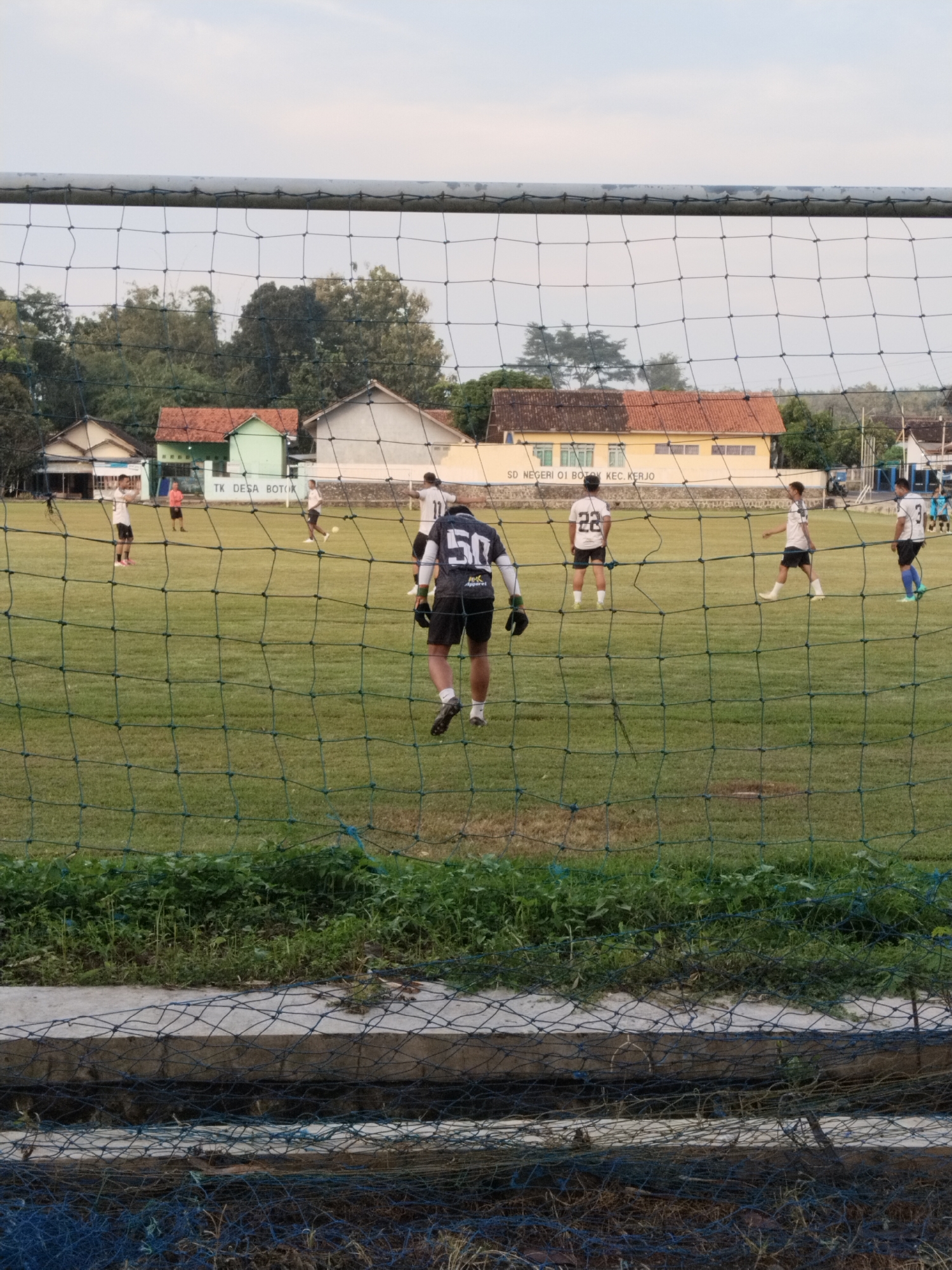 A low-angle outdoor shot, taken through a soccer goal net, shows a soccer field with players during what appears to be a training session or a relaxed game. Several players are visible on the field, wearing different colored jerseys with numbers on their backs, including "50" and "22." The players are spread out across the field, some standing, some walking.
In the background, there are buildings. The building with the sign “TK DESA BOTOK” is on the left side of the image and another building with the sign “SD NEGERI 01 BOTOK KEC. KERJO” is on the right. These signs provide location information: Botok, Indonesia. The background also includes trees and greenery.
The foreground is dominated by the soccer net, which is slightly out of focus, and some weeds and plants.