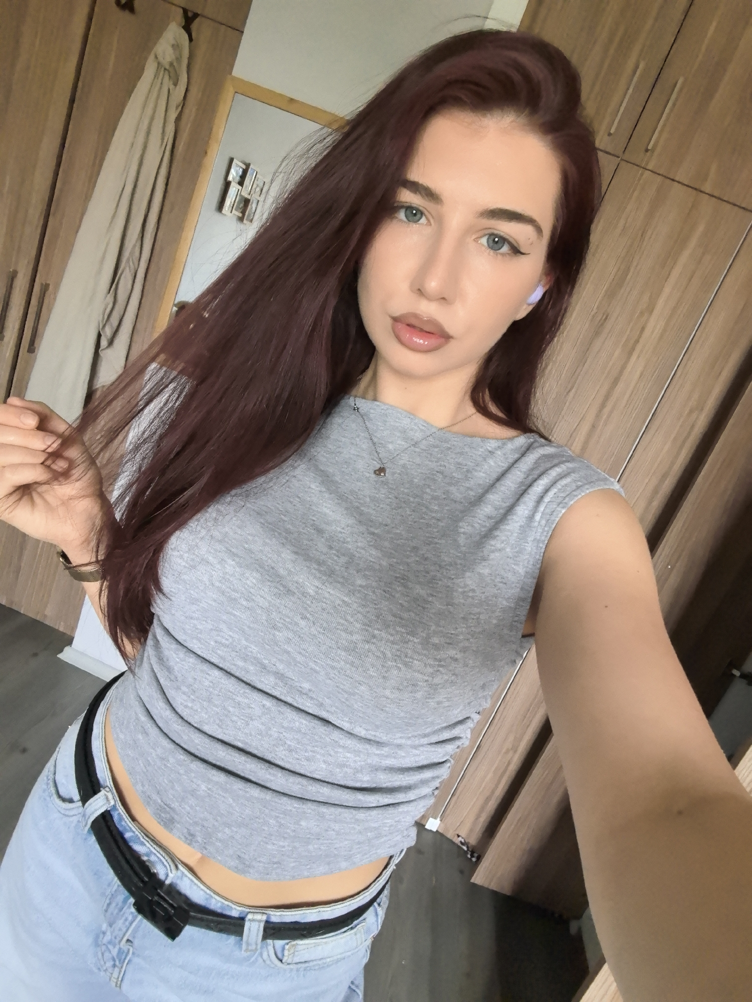 A young woman is captured in a selfie taken indoors, likely in a residential setting in Stara Zagora, Bulgaria. She appears to be in her early to mid-20s, featuring long, dark reddish-brown hair, light blue-green eyes, and fair skin. Her makeup includes winged eyeliner and glossy, nude-toned lipstick. A small, light purple earbud is visible in her left ear.
She is wearing a light grey, short-sleeved crop top with a boat neck, which is gathered around the waist, revealing her midriff. Her attire also includes light-wash blue jeans secured with a black belt. A silver necklace with a small heart-shaped pendant is visible around her neck, and a dark-colored watch or bracelet is on her left wrist.
The woman is looking directly at the camera with a neutral expression, gently holding a strand of her hair with her left hand. The background consists primarily of wooden wardrobes or closets, suggesting a bedroom or dressing area. On the left side, a light-colored garment hangs on a hook. A mirror reflects a portion of a white wall adorned with several small framed pictures or decorative items. A dark floor is partially visible at the bottom left of the frame. The lighting suggests it is daytime.