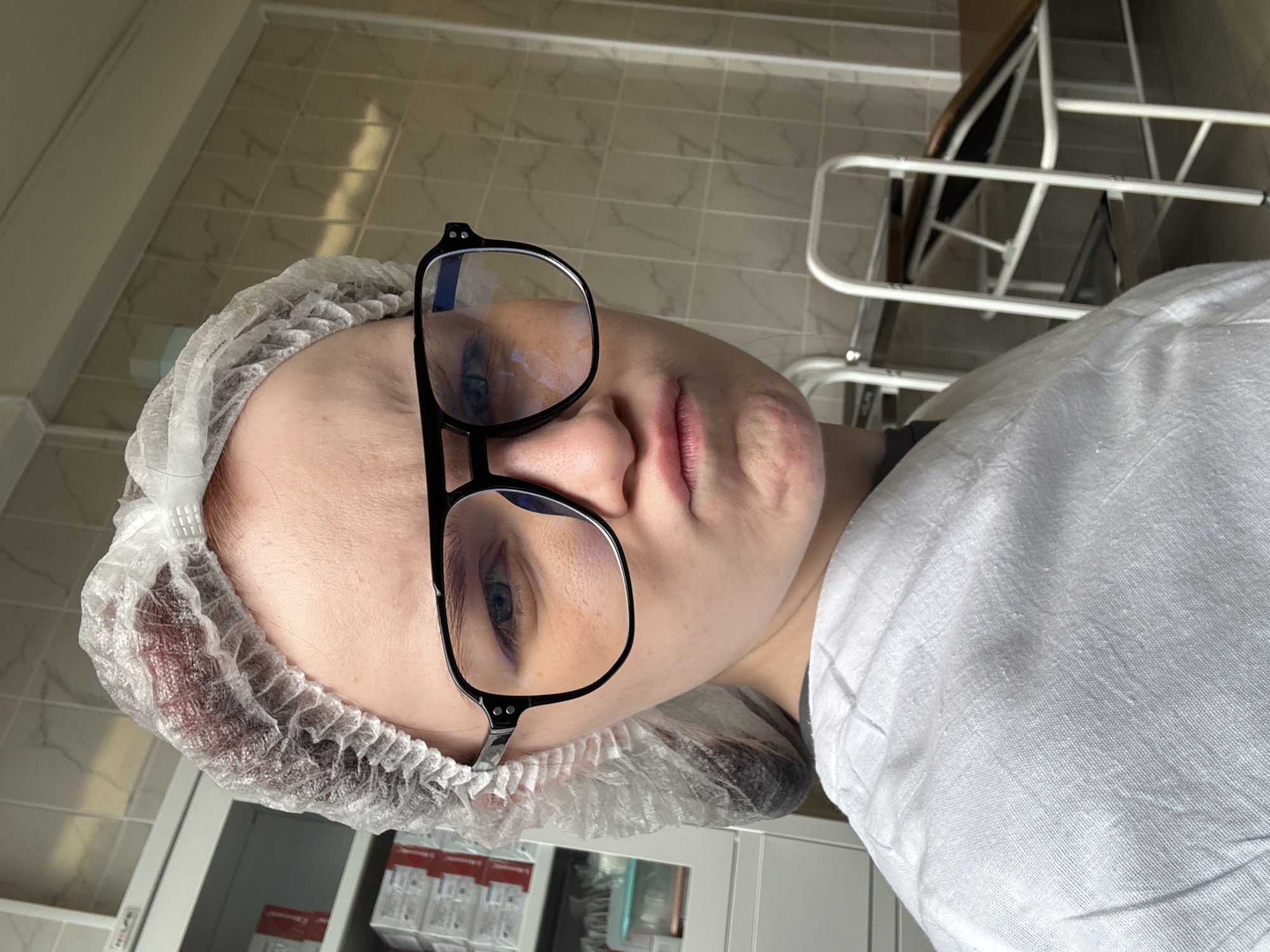 A young female, wearing a white disposable hairnet and glasses with black frames, pouts with pursed lips. She is dressed in a white medical gown or drape. The background appears to be a sterile environment, possibly a medical clinic or hospital room, with tiled walls and medical equipment such as shelves filled with boxes and an examination table visible. The lighting is neutral, and the overall impression is one of a casual selfie taken in a medical setting. The location is unknown.