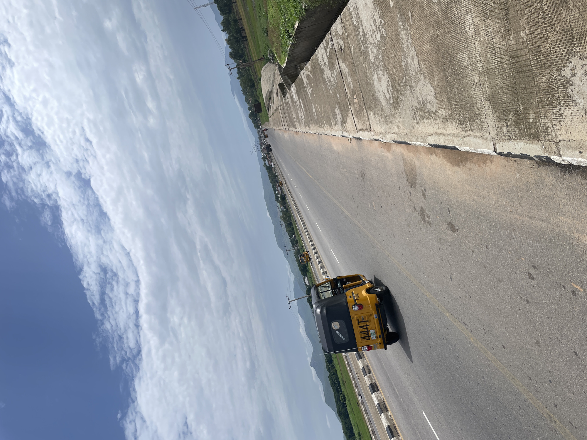 The image depicts a yellow and black auto-rickshaw driving on a highway in Jalingo, Nigeria. The auto-rickshaw is in the right lane, with its rear facing the viewer. The highway is a multi-lane road with a concrete barrier on the right side and a black and white striped shoulder. Green fields and trees are visible on the left side of the highway, with mountains in the background. The sky is cloudy and blue, suggesting it is daytime. The scene appears to be a rural or semi-rural area, given the natural landscape surrounding the highway. There is no discernible activity or event taking place beyond the auto-rickshaw traveling. The license plate on the auto-rickshaw displays the text "444T" and what appears to be a Nigerian license plate format. The overall mood of the image is neutral, conveying a sense of travel and everyday life in Nigeria.