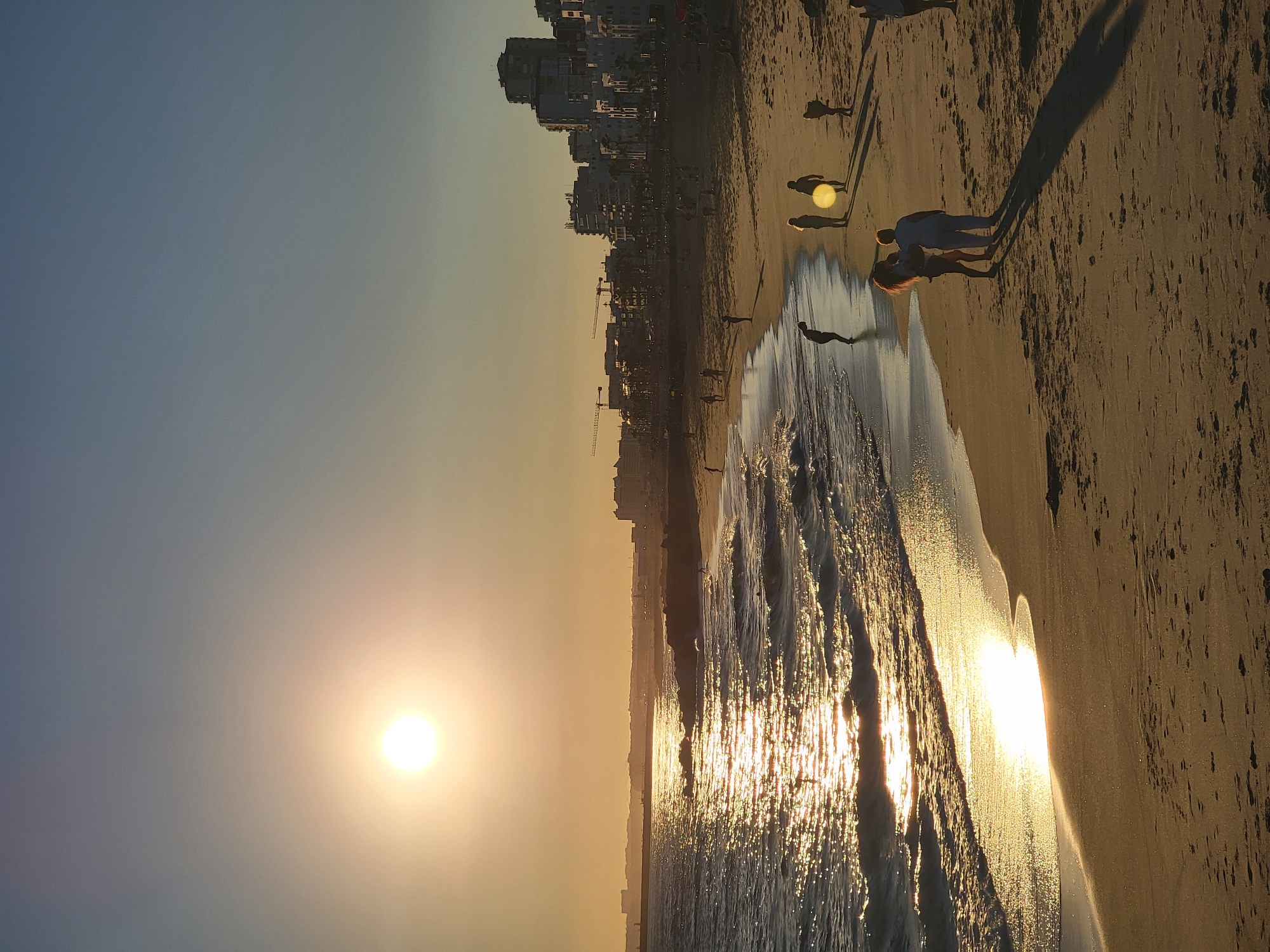 The image depicts a sunset scene at the beach in Quarteira, Portugal. The sun is low in the sky, casting a warm golden glow across the water and sand. Gentle waves are breaking on the shore, and the sunlight reflects off the water's surface, creating a shimmering effect. Several people are visible on the beach, some walking along the waterline, others standing and interacting. In the background, a line of buildings forms the Quarteira skyline, with construction cranes suggesting ongoing development. The atmosphere is serene and peaceful, characteristic of an evening at the coast.