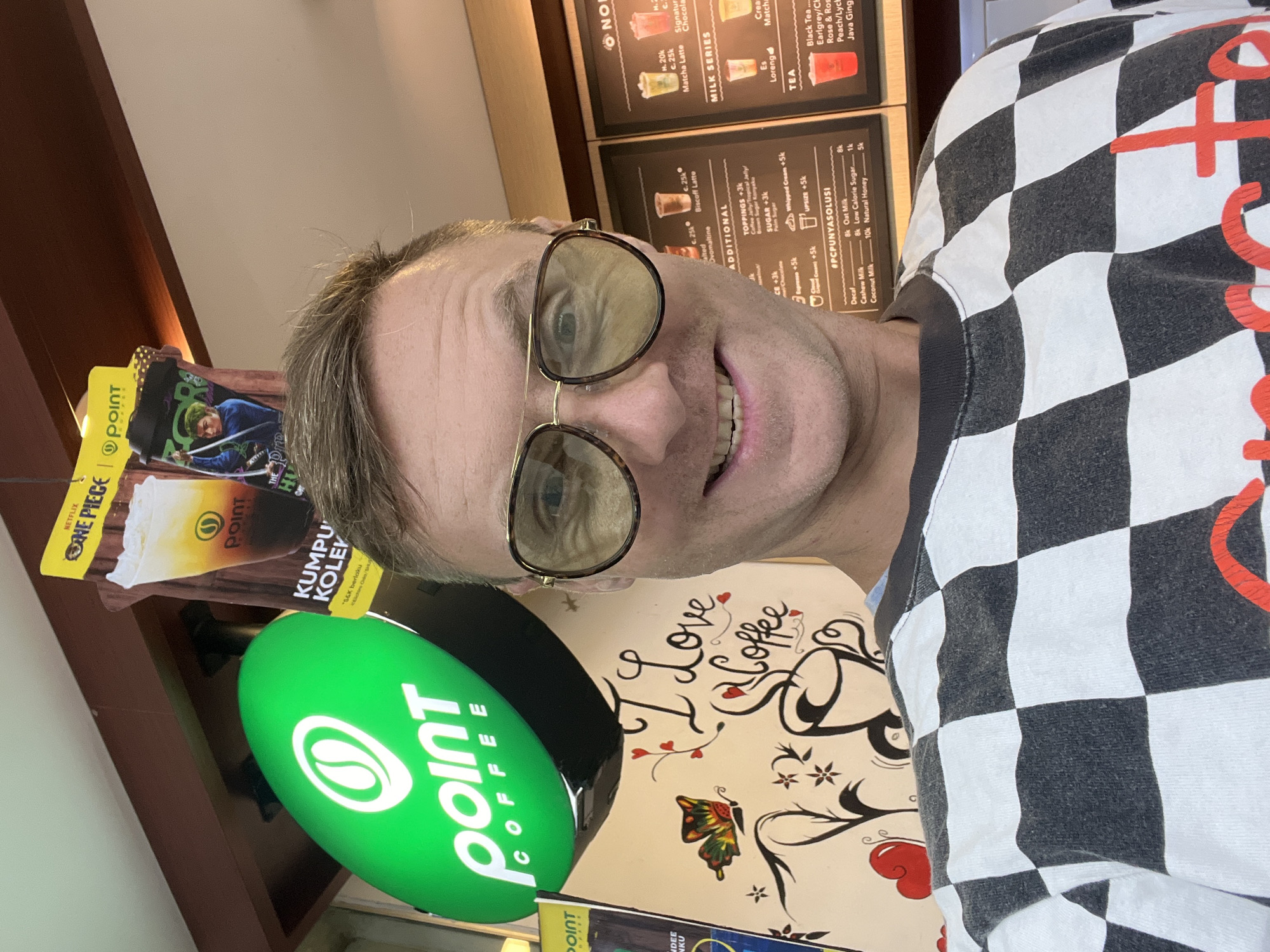 A man is taking a selfie in front of a Point Coffee shop in Sukawati, Indonesia. The man is wearing a black and white checkered long-sleeved shirt and large aviator-style sunglasses. In the background, a green oval sign with a white coffee bean logo and the words "point COFFEE" is visible. There are also promotional posters for drinks, one featuring characters from the anime series "One Piece." A wall behind the man is decorated with a hand-drawn mural that includes the words "I Love Coffee," a coffee cup, and a butterfly. The man appears to be smiling. The environment suggests an indoor retail space, likely a coffee shop.
