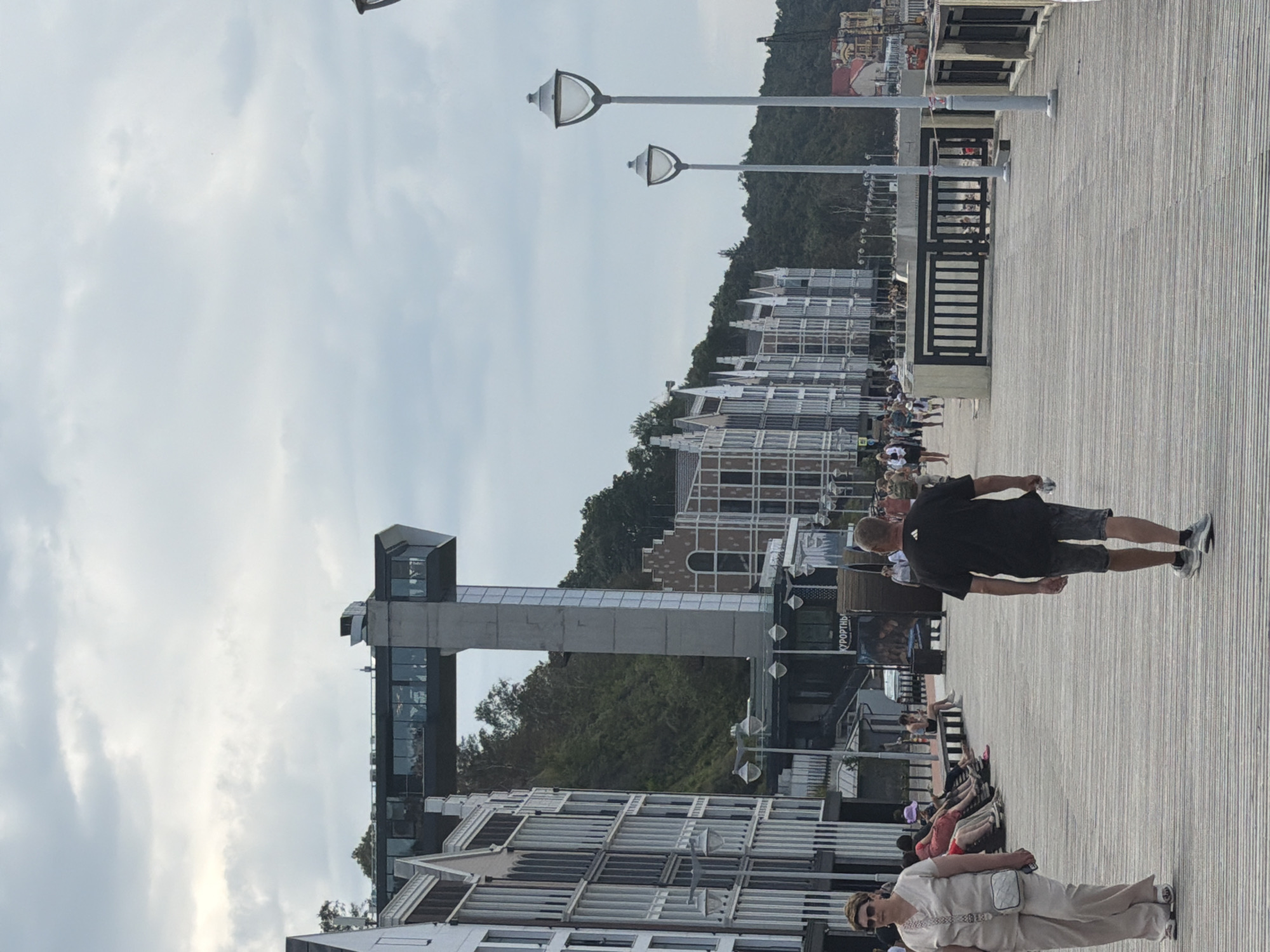 The image depicts a wide wooden boardwalk with a textured surface in Svetlogorsk, Russia. The boardwalk is lined with lampposts, and on the left, there are modern buildings with a European architectural style. Further in the background, a prominent structure resembling a futuristic observation deck or elevator tower rises from a forested hillside. A few people are scattered on the boardwalk, some walking and others sitting on benches. A woman in a light-colored outfit and a man in a black t-shirt and shorts are walking away from the camera. The sky is overcast with scattered clouds.