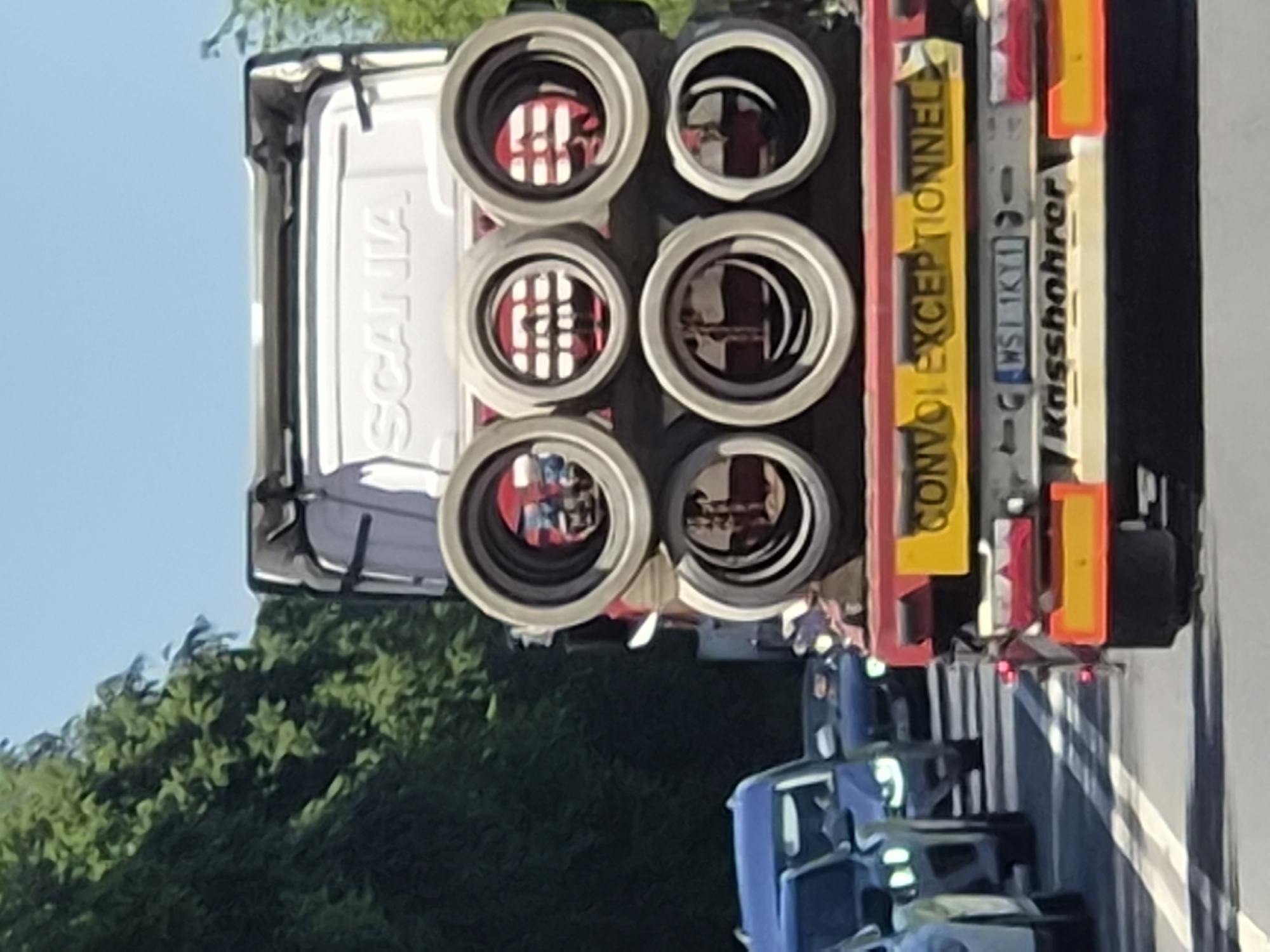 The image shows a white Scania truck, possibly a heavy hauler, with six round lights mounted on its back. The lights reflect red and white patterns. A yellow sign reads "CONVOI EXCEPTIONNEL" indicating an oversized load. The license plate, partially visible, reads "WSI 1KY11" and is German. The text "Kassbohrer" is also visible, likely indicating the manufacturer.
Based on the license plate and signage, the truck is in Luxembourg, most likely traveling on a road based on the road markings and presence of other vehicles, as well as the trees in the background.