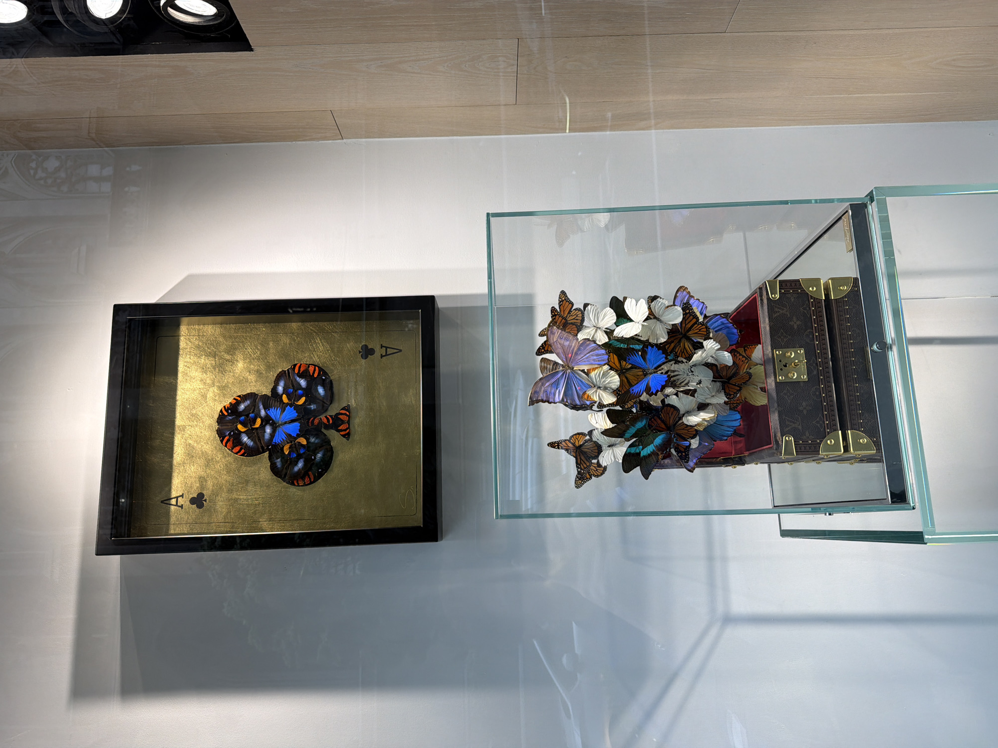 The image shows a display of artwork on a wall, likely indoors. On the left is a framed piece depicting an Ace of Clubs playing card, with a golden background and what appears to be a collage of butterfly wings forming the club symbol. To the right is a clear glass box containing a miniature trunk and a collection of artificial flowers and colorful butterfly models. The lighting is bright and the background is a neutral color, suggesting a gallery or exhibition space. Without further information, determining the exact location within New York City is not possible, though the overall ambiance suggests a high-end or artistic setting.