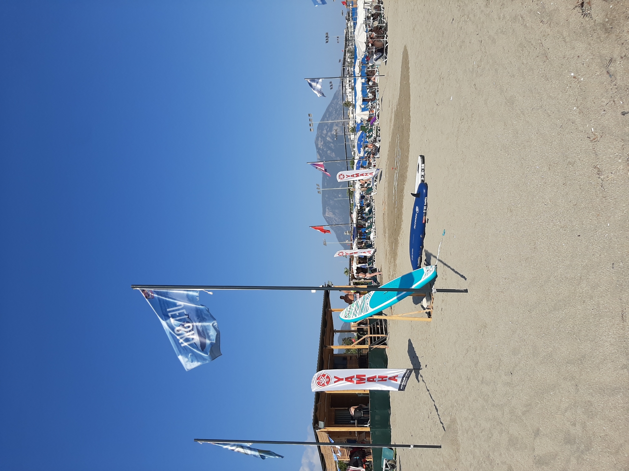 The image captures a beach scene in Alanya, Turkey, under a clear blue sky. In the foreground, the sandy beach is populated with several surfboards and paddleboards, some standing upright on wooden stands. A prominent Yamaha flag, a white banner with red lettering and a logo, stands vertically in the sand. To the left, another flag, light blue with white graphics, is partially visible. In the mid-ground, a beachside structure with a wooden exterior and a thatched roof serves as a cafe or rental shop, with people seated inside and on its steps. Further back, rows of white beach umbrellas and lounge chairs indicate a recreational area, with more people relaxing. In the distance, a large, mountainous landscape is visible, with streetlights and some buildings dotted along the coastline. The overall atmosphere is one of a sunny day at a popular tourist destination.