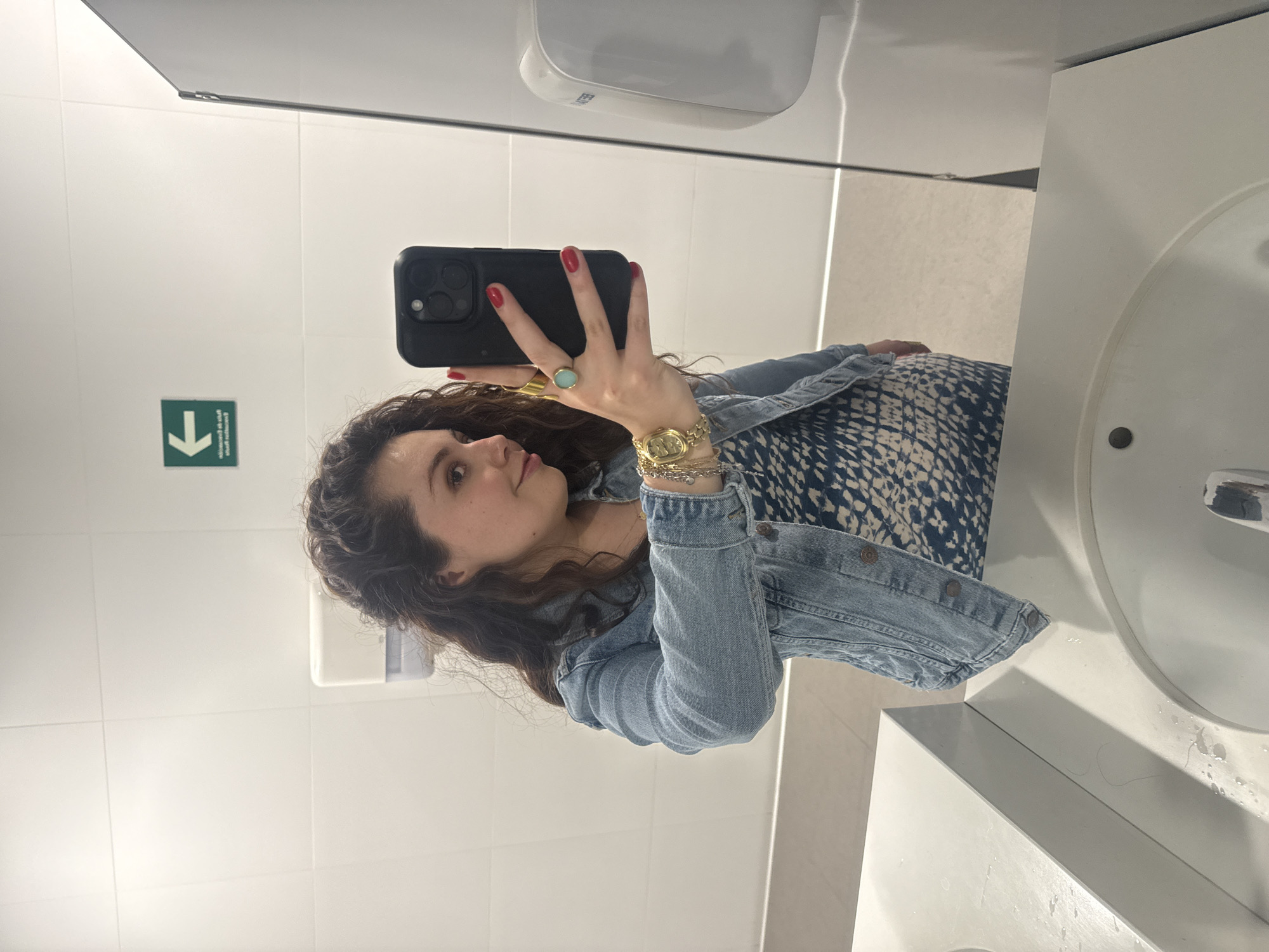 A woman is taking a selfie in a bathroom. She is holding a black iPhone in her right hand, which is adorned with a turquoise ring and gold jewelry. The woman has curly brown hair and is wearing a denim jacket over a blue and white patterned dress. The background of the image shows white tiled walls, a sink with a silver faucet, a hand dryer, and an emergency exit sign with a downward-pointing arrow. Given the overall context and style, it is not possible to pinpoint the exact location within Mexico City.