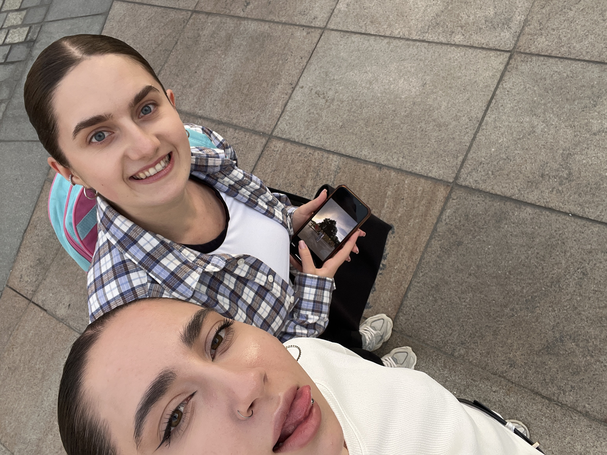 Two young women are taking a selfie on a paved walkway. The woman in the foreground, on the left, is wearing a white ribbed top and has her tongue out, with a piercing on her tongue. She has dark hair pulled back and is wearing a nose ring and hoop earrings. The woman behind her, on the right, is wearing a blue and white plaid shirt over a white top, and a pink and blue backpack. She has dark hair pulled back and is smiling, showing her teeth. She is holding a smartphone in her hands. The ground is paved with large, square tiles in various shades of gray and beige. The image is taken outdoors in what appears to be an urban setting. The setting is consistent with areas in the city of Lublin, Poland, such as pedestrian zones or park pathways.