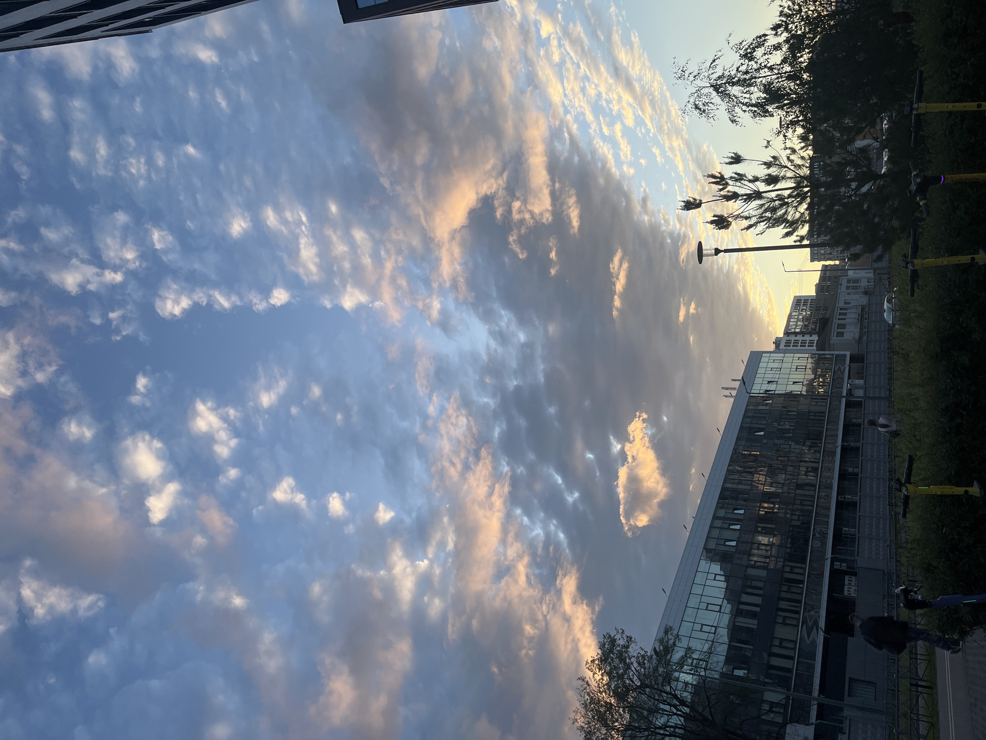 This is an image of a modern building with reflective windows in Saint Petersburg, Russia, during what appears to be either sunrise or sunset. The sky is filled with a mix of gray and pinkish clouds, with patches of blue visible. In the foreground, there is a paved walkway, a line of green bushes, and several electric scooters parked along the path. Two people are walking on the path, and a third person is standing near the bushes. The building's facade reflects the sky, creating a mirror-like effect. There are trees and streetlights visible in the scene, adding to the urban environment. The overall atmosphere is calm and serene, with the soft light of the sky illuminating the scene.