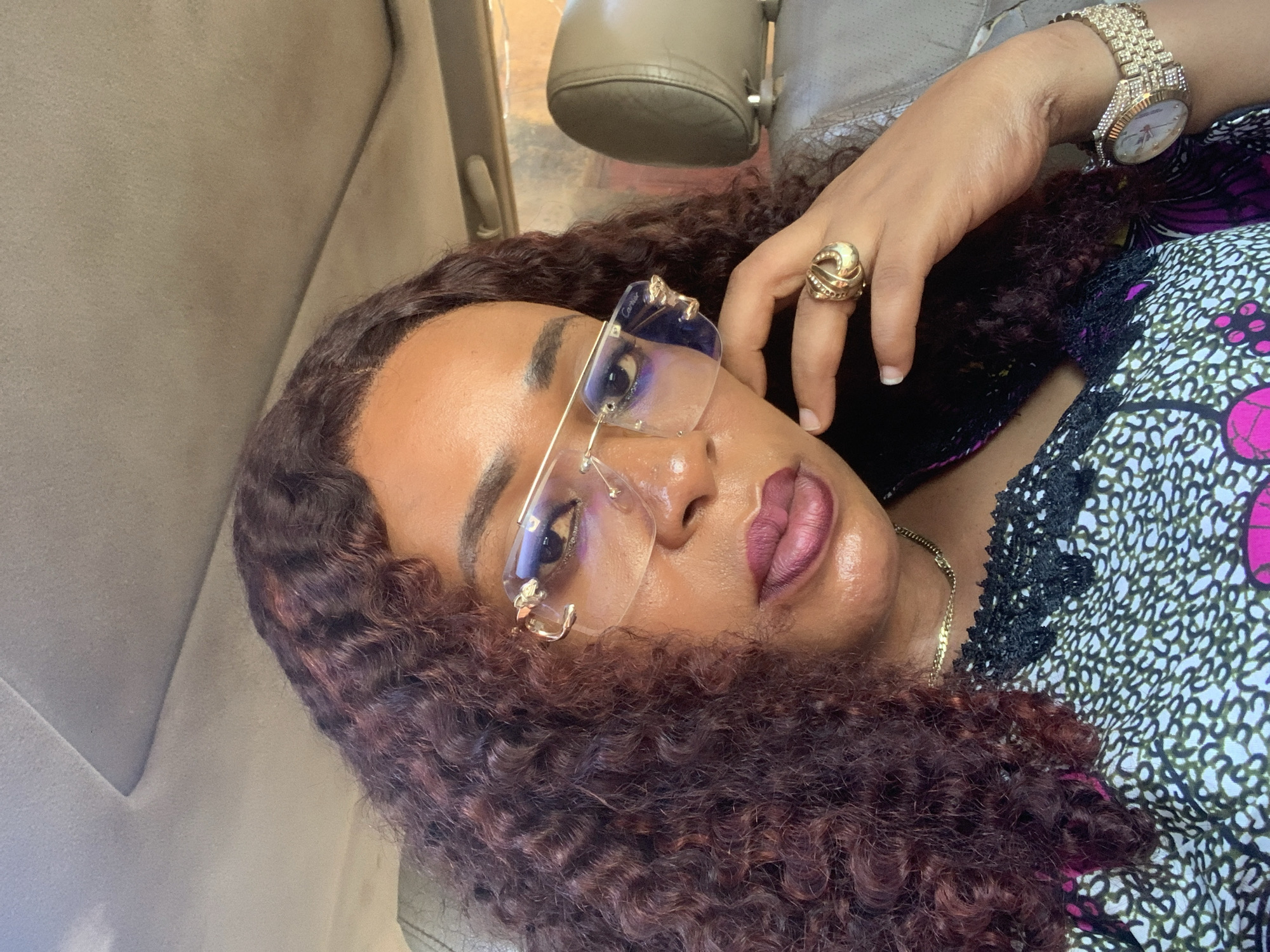 A woman is depicted in a close-up, self-portrait style image, situated within the interior of a vehicle in Sabon Tasha, Nigeria. She has a medium-dark complexion and long, dark reddish-brown curly hair. Her lips are adorned with a plum-colored lipstick that has a glossy finish, and her eyebrows are dark and well-defined. She is wearing rimless eyeglasses with gold-toned hardware, which includes a detail resembling a panther head on the hinge and the word "Cartier" visible on the lens in a cropped view. A large, gold-toned, sculptural ring is on her right ring finger, and a gold-toned wristwatch with a band encrusted with clear stones is on her left wrist. A thin, gold-toned chain necklace is visible at her neckline. Her top features an abstract pattern in white, black, olive green, and pink, and has a black lace trim along the neckline. Her right hand is raised, with fingers lightly touching her jawline. The vehicle's interior headliner is light-colored, and a light-colored car seat with a headrest is visible in the background to the right. Natural light illuminates the scene.