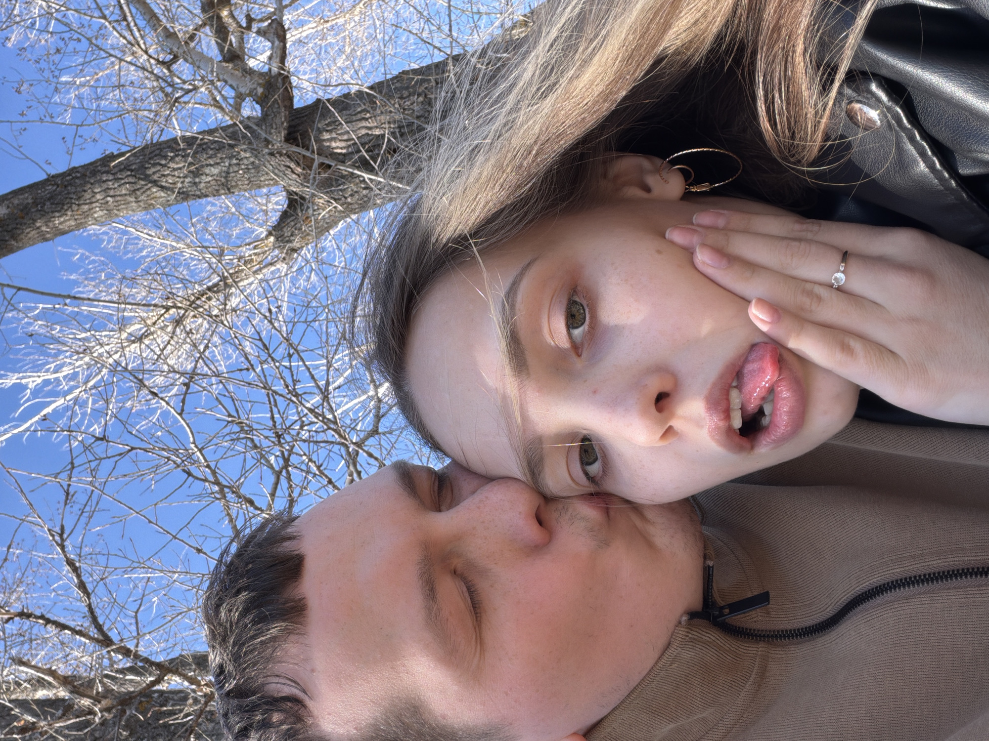 A young couple poses for a selfie against a backdrop of a bare tree and a bright blue sky. The woman, with light blonde hair, wears a black leather jacket and gold hoop earrings, displays a ring on her finger, and sticks out her tongue. The man, with dark hair, has his eyes closed and appears to be kissing the woman. The photo was taken outdoors, possibly in the city of Chaikovsky, Russia, as specified in the prompt.