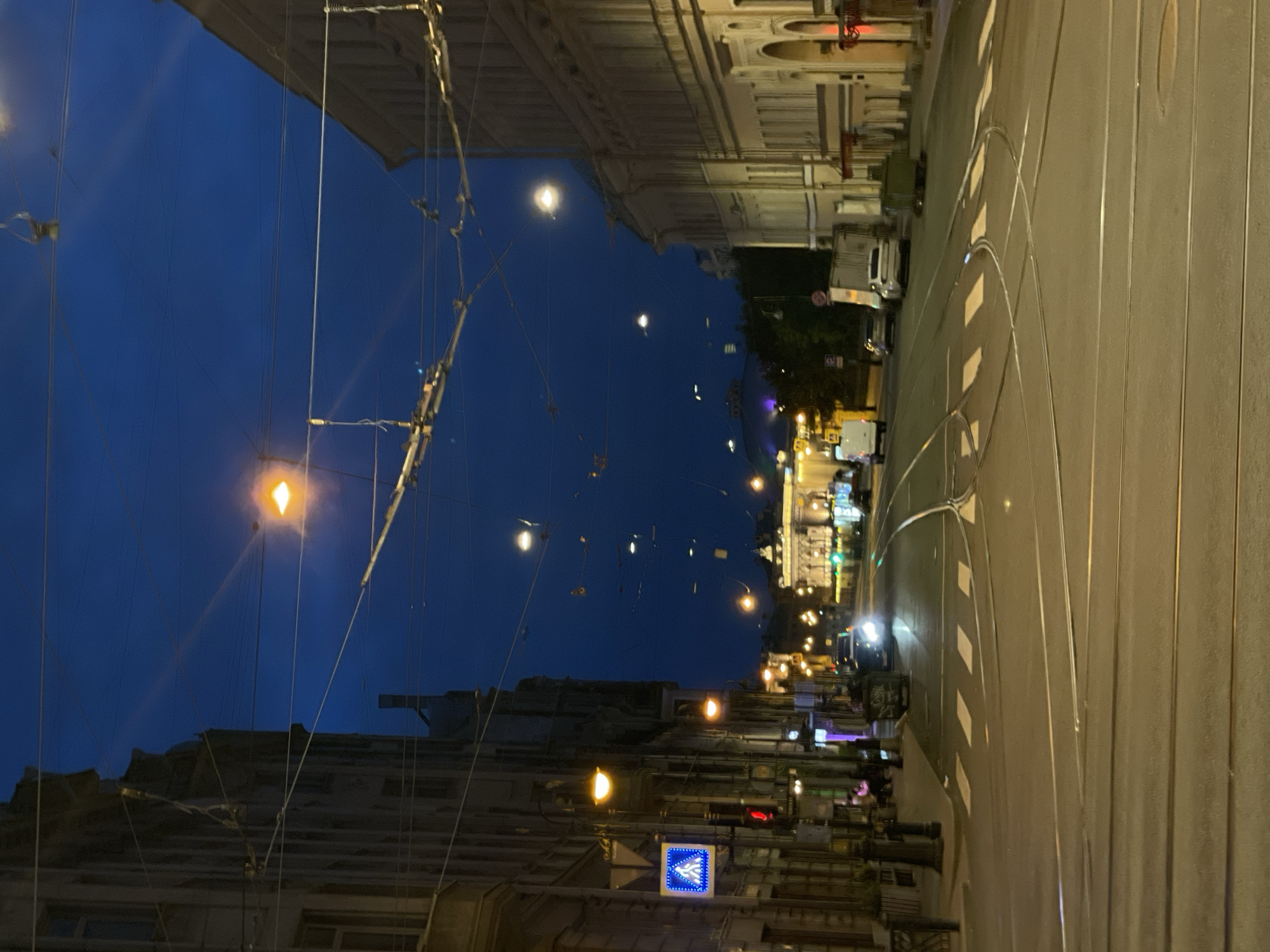 The image captures a nighttime view of a city street in Saint Petersburg, Russia, likely during the twilight hours due to the dark blue sky illuminated by streetlights. The scene is dominated by a wide asphalt road with multiple sets of tram tracks, indicating a well-trafficked urban area. On either side of the street are substantial, multi-story buildings with classical architectural features, typical of Saint Petersburg.
The buildings on the left have a darker, more imposing facade, while those on the right are lighter in color and appear more ornate. Numerous overhead wires, characteristic of a tram or trolleybus system, crisscross the sky, supported by poles and structures. Several streetlights are visible, casting a warm, orange glow on the road and sidewalks.
In the foreground, the tram tracks form a complex junction, suggesting a major intersection. Pedestrian crossings are marked with white stripes on the road. A pedestrian crossing sign with a blue background and a white illuminated pedestrian symbol is mounted on a pole on the left side, along with a red traffic light.
Several vehicles are present on the street. A white van is seen in the distance on the left, and a car with its headlights on is approaching from the right. Other cars are parked or driving further down the road. A green dumpster is visible on the right side of the street. A few figures are seated on a bench outside a building on the left, suggesting the presence of cafes or shops. In the far distance, a grand illuminated building with classical architecture stands out, possibly a landmark. The overall impression is of a vibrant city street at dusk, a blend of historical architecture and modern urban infrastructure.