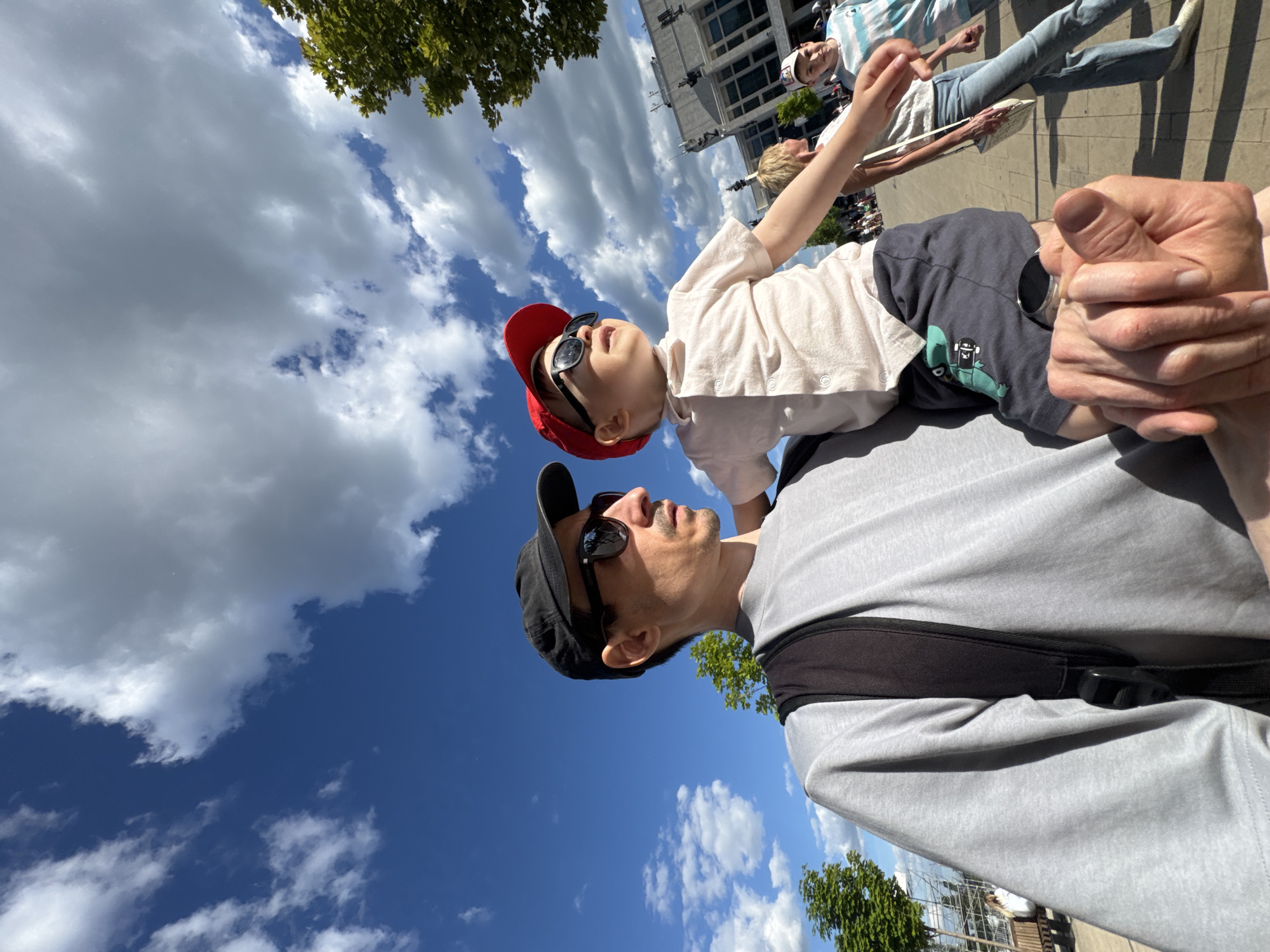 A low-angle, wide-perspective photograph captures a man carrying a small child in a brightly lit outdoor urban environment under a blue sky with scattered cumulus clouds. The scene is located in Izhevsk, Russia, in the vicinity of the Izhevsk State Opera and Ballet Theatre, identifiable by the distinctive architectural building in the mid-ground right.
In the foreground, a man, viewed from the chest up and slightly to his left, wears a grey short-sleeved t-shirt, a black baseball cap, and dark sunglasses. He has a slight beard and mustache and is looking towards the right side of the frame. A black backpack strap is visible on his right shoulder. His right hand, positioned in the bottom right corner, supports the child.
The small child, held at the man's hip or shoulder level, is positioned to the right of the man and looks upward and to the right, also wearing dark sunglasses and a red baseball cap. The child wears a light-colored, collared short-sleeved shirt with buttons down the front and dark shorts featuring a green dinosaur graphic on the left leg.
The background reveals a paved pedestrian area with several blurred individuals walking. To the far right, a woman in light blue jeans and a light-colored top carries a white shoulder bag, walking to the left. A young boy in a striped t-shirt and a light-colored baseball cap walks beside her. Green-leafed trees are present on both the left and right sides of the frame. The sunlight casts distinct shadows, indicating a bright day.