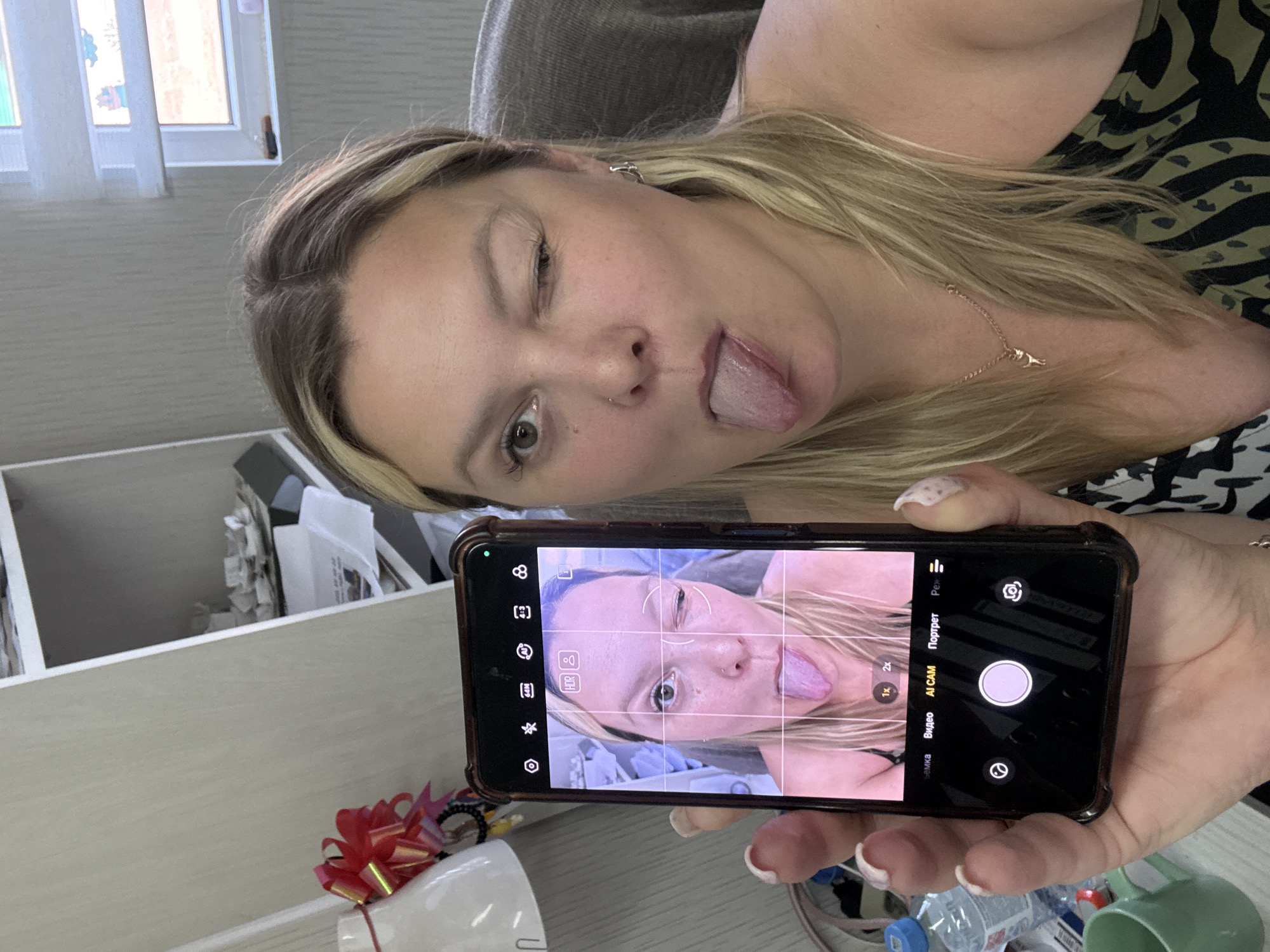 A woman with blonde hair and light skin is holding a smartphone in her left hand, taking a selfie. She is winking and sticking her tongue out. Her right eye is closed, and her tongue is extended. The smartphone screen shows a mirrored image of her face, also with her winking and sticking her tongue out. The phone is held in a way that the top of the phone is facing the viewer. She is wearing a necklace with a small pendant. The background appears to be an indoor setting, possibly an office or a room with shelves in the back. The lighting suggests it is daytime. The image does not provide any specific information about the location in the city of Noviy, Russia.