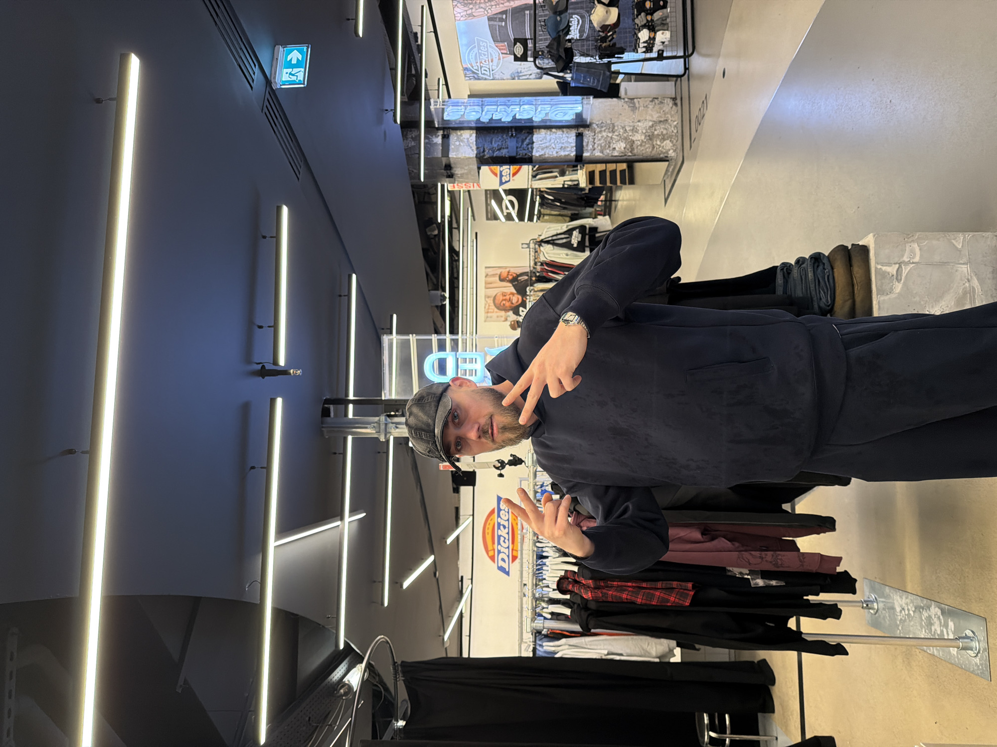 A man wearing a gray hat and a navy blue sweatsuit is standing inside a Dickies store. The Dickies logo is visible behind him. He is making a peace sign with both hands. Clothing racks with various garments are in the background. The store's interior features modern lighting. The image appears to have been taken indoors at a retail location, potentially a Dickies store, in Paris, France.