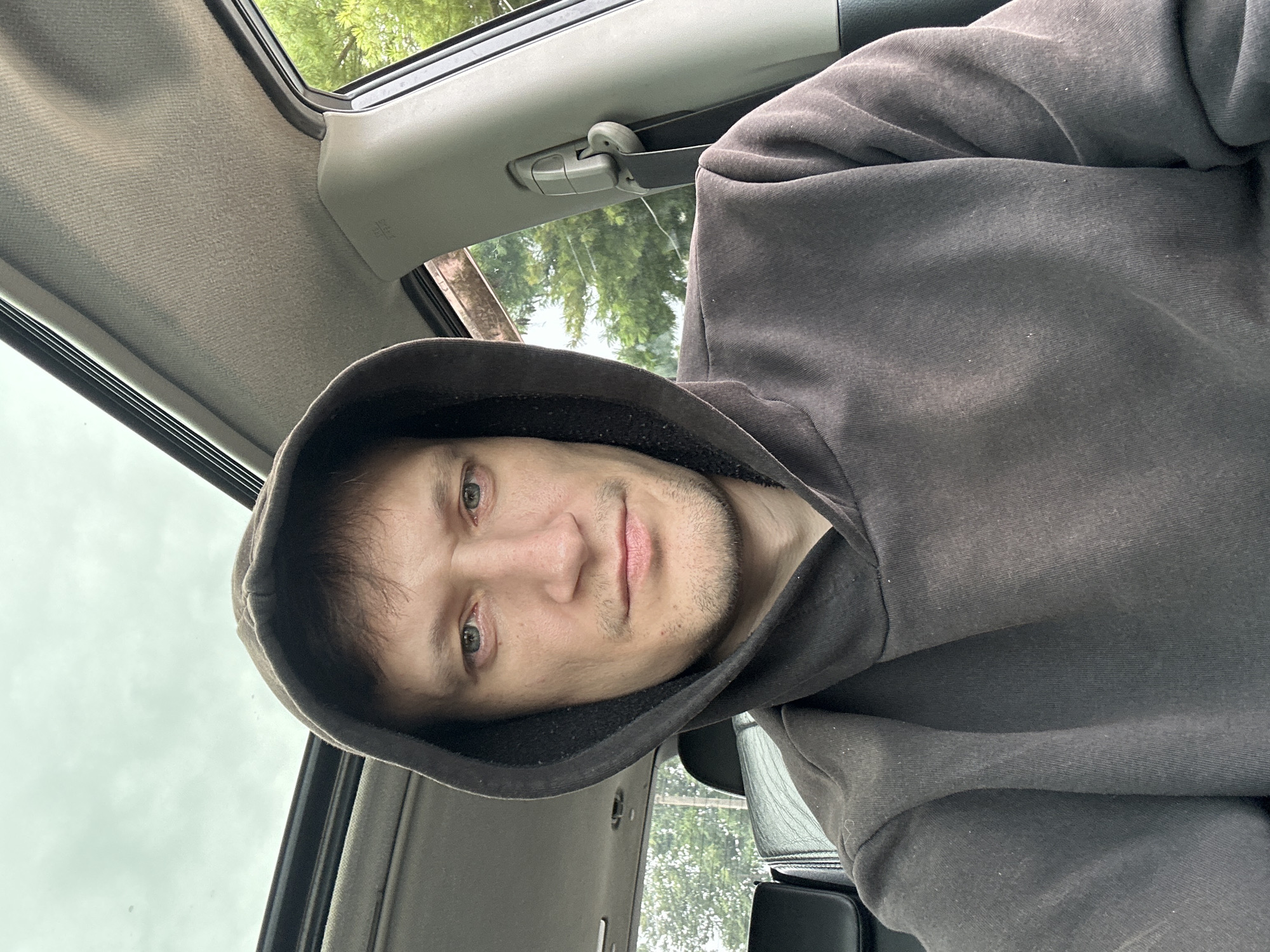A medium shot shows a man in a dark grey hoodie inside a car. He is looking directly at the camera with a neutral expression. His eyes are open, and his mouth is slightly closed. The man has light skin and short brown hair. The car's interior is visible, including the ceiling with a sunroof, a portion of the driver's seat, and the passenger seat headrest. Through the window, trees and foliage can be seen, suggesting an outdoor, possibly suburban or rural, setting. The sky appears overcast. The image is taken from a selfie perspective.