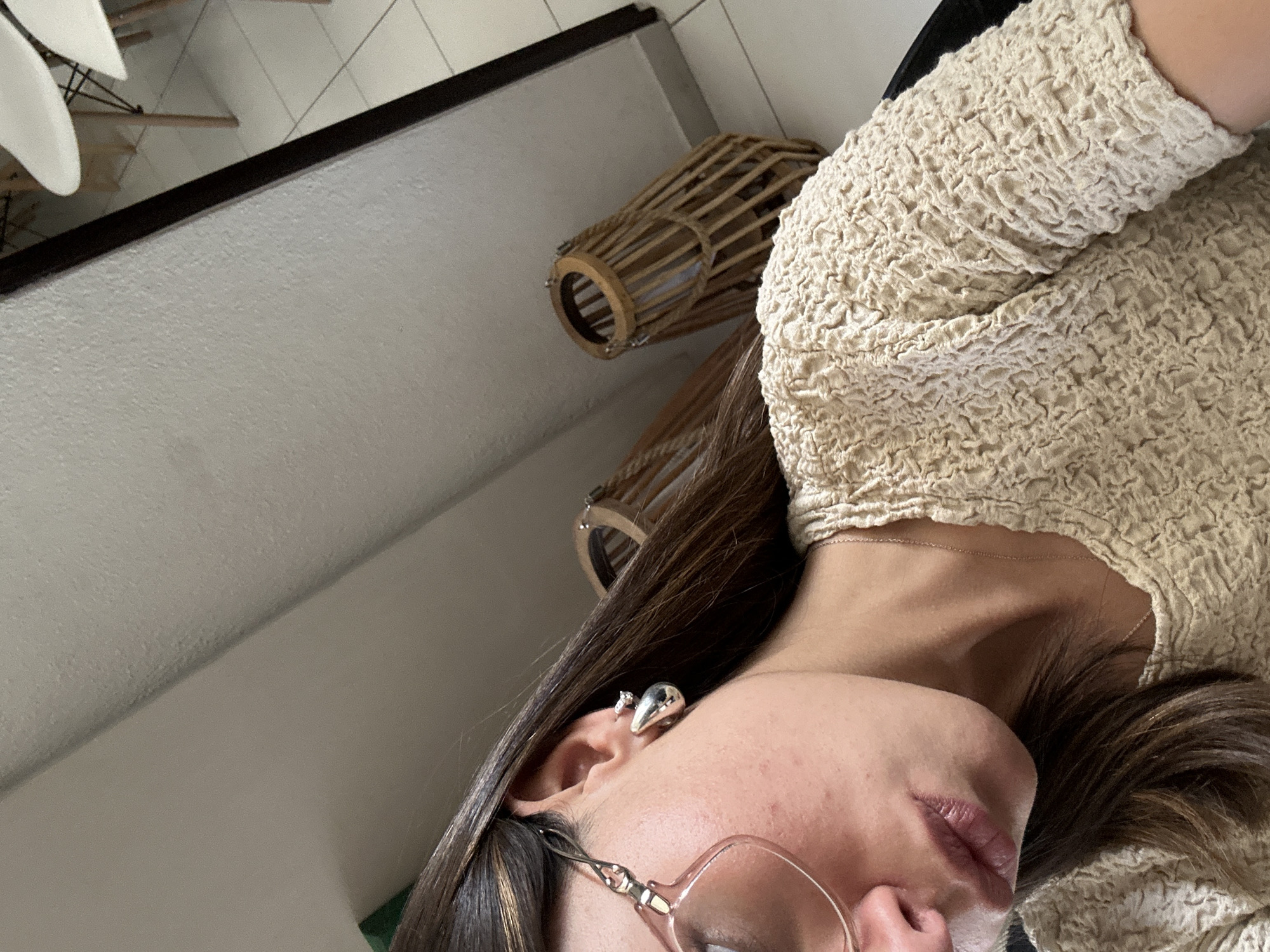 The image shows a close-up of a woman's face and torso, seemingly indoors. She has brown hair and is wearing glasses and silver earrings. She is wearing a beige, textured top. In the background, there is a white wall, a dark ledge or frame, and two wicker-like objects that appear to be hanging or placed on a surface. There are also hints of a white chair in the upper left corner. The location is Unknown.