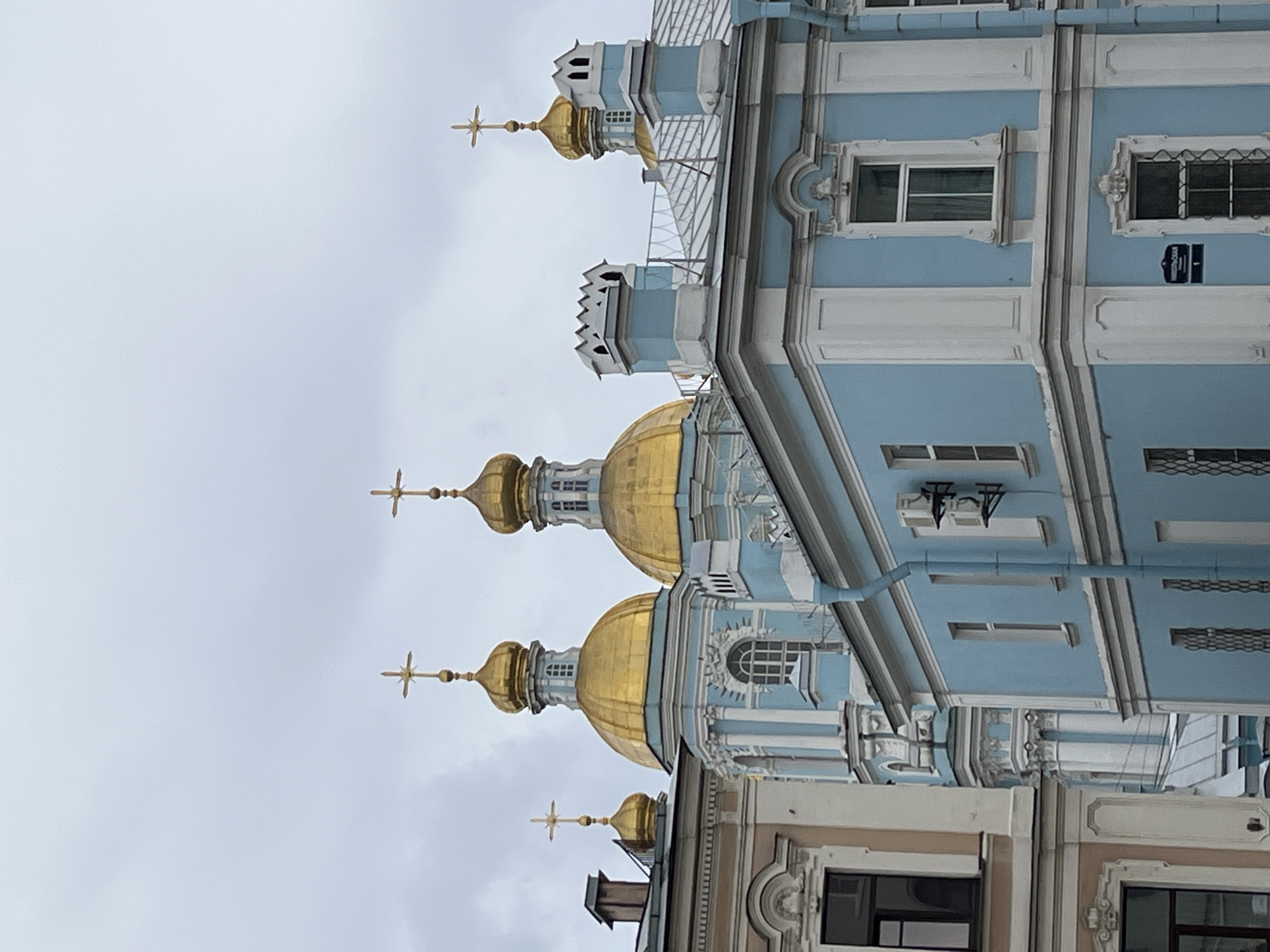 The image shows a vertical, low-angle view of a building, identified as the Naval Cathedral of Saint Nicholas in Saint Petersburg, Russia. The building features a light blue facade with white trim. Several golden domes topped with crosses are visible. The sky is overcast. A street sign is attached to the side of the building. No people are visible in the image.