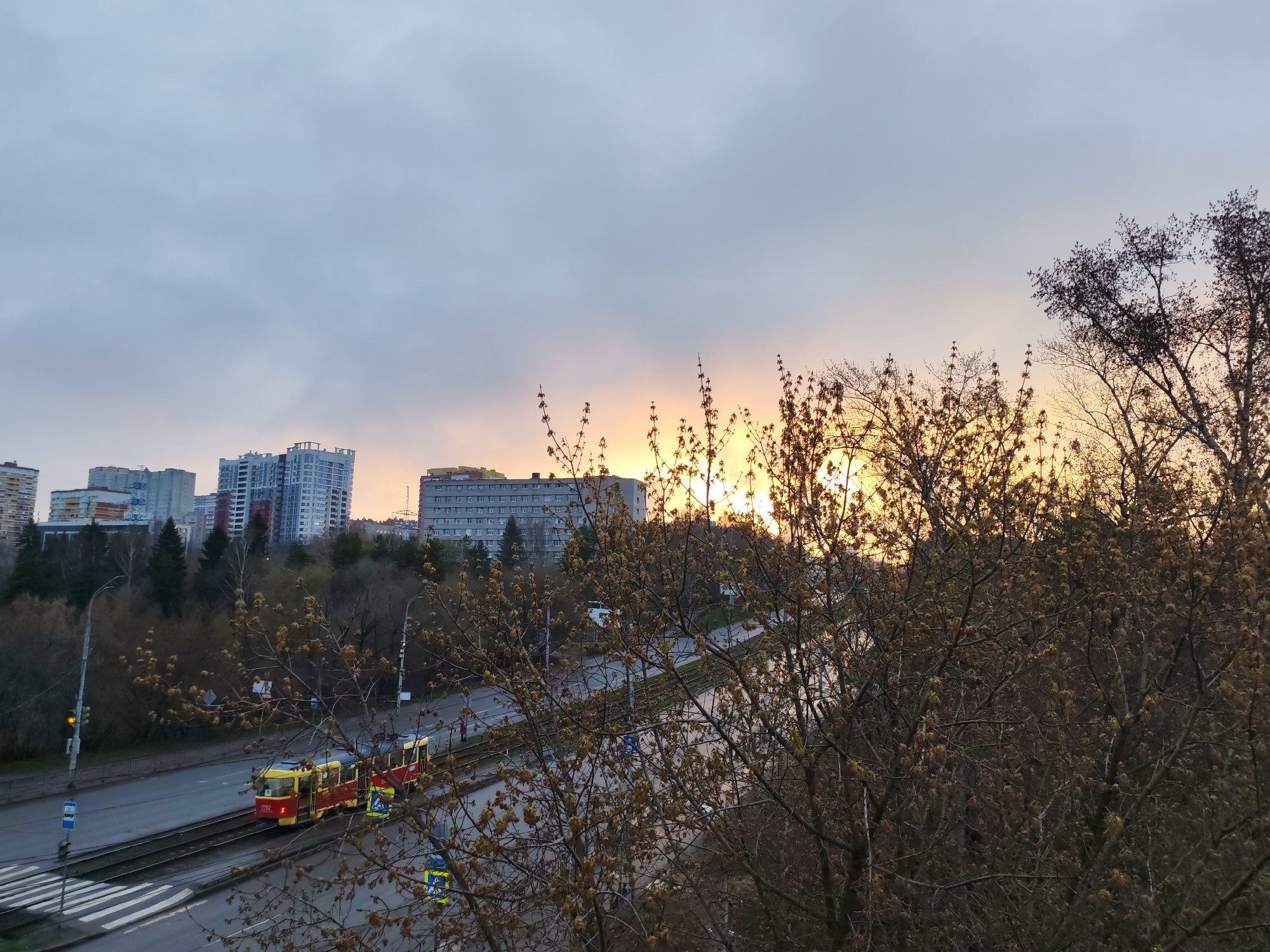 The image shows a cityscape during sunrise/sunset in Izhevsk, Russia. In the foreground, bare trees with budding leaves partially obscure the view of a street with tram tracks. A red and yellow tram is visible on the tracks, heading towards the viewer's left. The street also contains vehicle traffic. In the background, various buildings and apartment complexes of varying heights are visible, with the skyline silhouetted against the bright light of the rising or setting sun. The sky is cloudy with colors ranging from grey to orange and yellow near the horizon. Pedestrian crossing, street signs, and poles can be seen along the road.