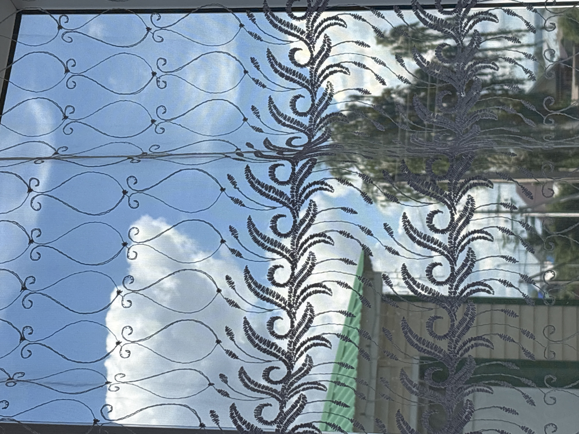 The image shows a sheer curtain with an intricate, dark grey embroidered floral pattern, viewed from indoors. Through the curtain, a bright blue sky with scattered white clouds is visible. The curtain is framed by a white window frame on the right side.
Below the sky, the pattern of the curtain changes to a repeating design of swirling, fern-like fronds. Through this lower portion of the curtain, a building with a light-colored, horizontal siding and a green roof is visible. The building appears to be one or two stories high. Behind the building, trees with green foliage can be seen, suggesting an outdoor environment.
The scene is likely set in a residential area. Given the specific location of Alexeyevskoye, Russia, this could be a view from a home, an apartment building, or a public building with a window overlooking a yard or street. The light suggests it is daytime, likely sunny given the brightness of the sky and clouds. There are no people or animals clearly depicted in the image.