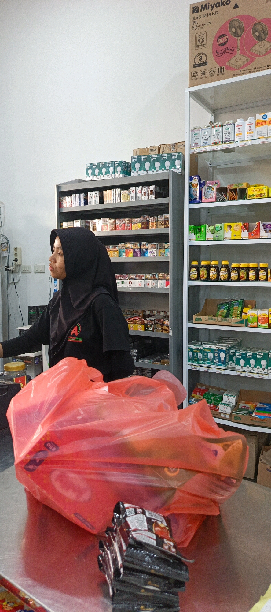 The image depicts an interior scene, likely a small retail establishment such as a minimarket or warung, located in Desa Simpang Katis, Indonesia. The foreground is dominated by a large, crinkled red plastic bag, possibly containing purchased items, and a stack of small, dark-packaged goods, likely snacks or confectionery, placed on a reflective surface.
In the mid-ground, a woman wearing a black hijab and a black t-shirt with a red and green logo that reads "A ABRAHAMMART" is partially visible, facing left. She appears to be attending to a customer or operating the establishment. Behind her, shelves are densely stocked with a variety of products. The shelves on the left are filled with numerous cigarette packs, arranged in rows. The shelves on the right display a diverse assortment of packaged goods, including boxes of light bulbs, containers of what appear to be traditional medicines or supplements (some labeled "HEROCYN" and "PRICKLY HEAT"), and rows of small amber-colored bottles, potentially containing beverages or health tonics. Further up on the right, there are boxes of various consumer products.
The environment appears to be brightly lit, with white walls visible in the upper left portion of the frame. Electrical outlets are mounted on the wall to the left of the woman. The overall impression is of a functional retail space stocked with a wide range of everyday items.
