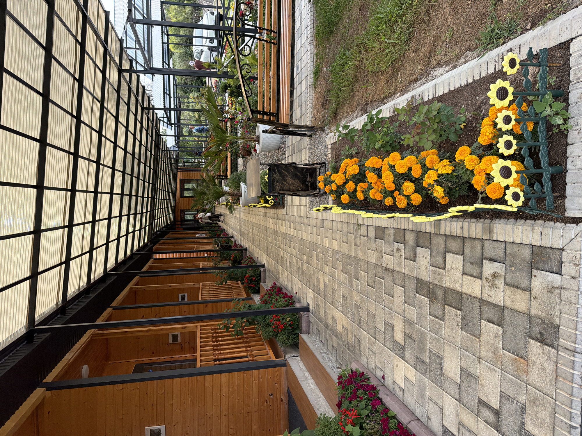 The image shows a long, covered walkway with wooden cabins on the left and a garden area with benches and plants on the right. The walkway is paved with light-colored square tiles. The cabins appear to be made of wood and have a porch area with railings. The garden area features various plants, including orange marigolds and decorative yellow sunflowers, as well as a wooden bench swing. In the background, there are several people walking and a white car parked. The scene suggests an outdoor recreational or lodging area.
