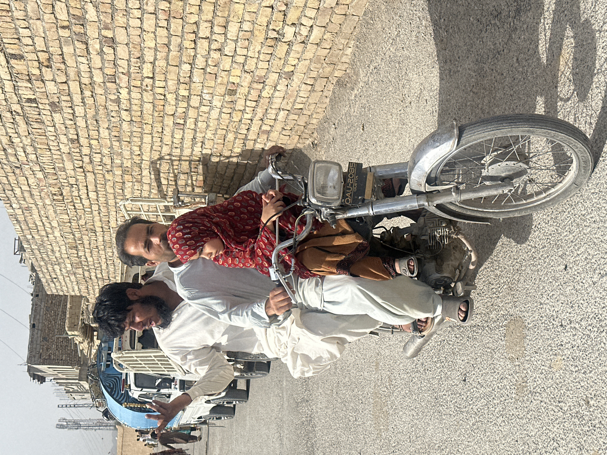 In Quetta City Tehsil, Pakistan, a man with a dark beard and wearing a light-colored robe gestures with his left hand while sitting on a motorcycle. Behind him, another man with short dark hair and light clothing supports a child in a red patterned dress and tan pants. The motorcycle, bearing the license plate "QAU-2085 QUETTA," is on a light-colored asphalt surface. Shadows are cast on the ground. In the background are buildings with light-colored brick and various vehicles, including a blue and white decorated auto-rickshaw, a white van, and other cars. The sky is overcast.