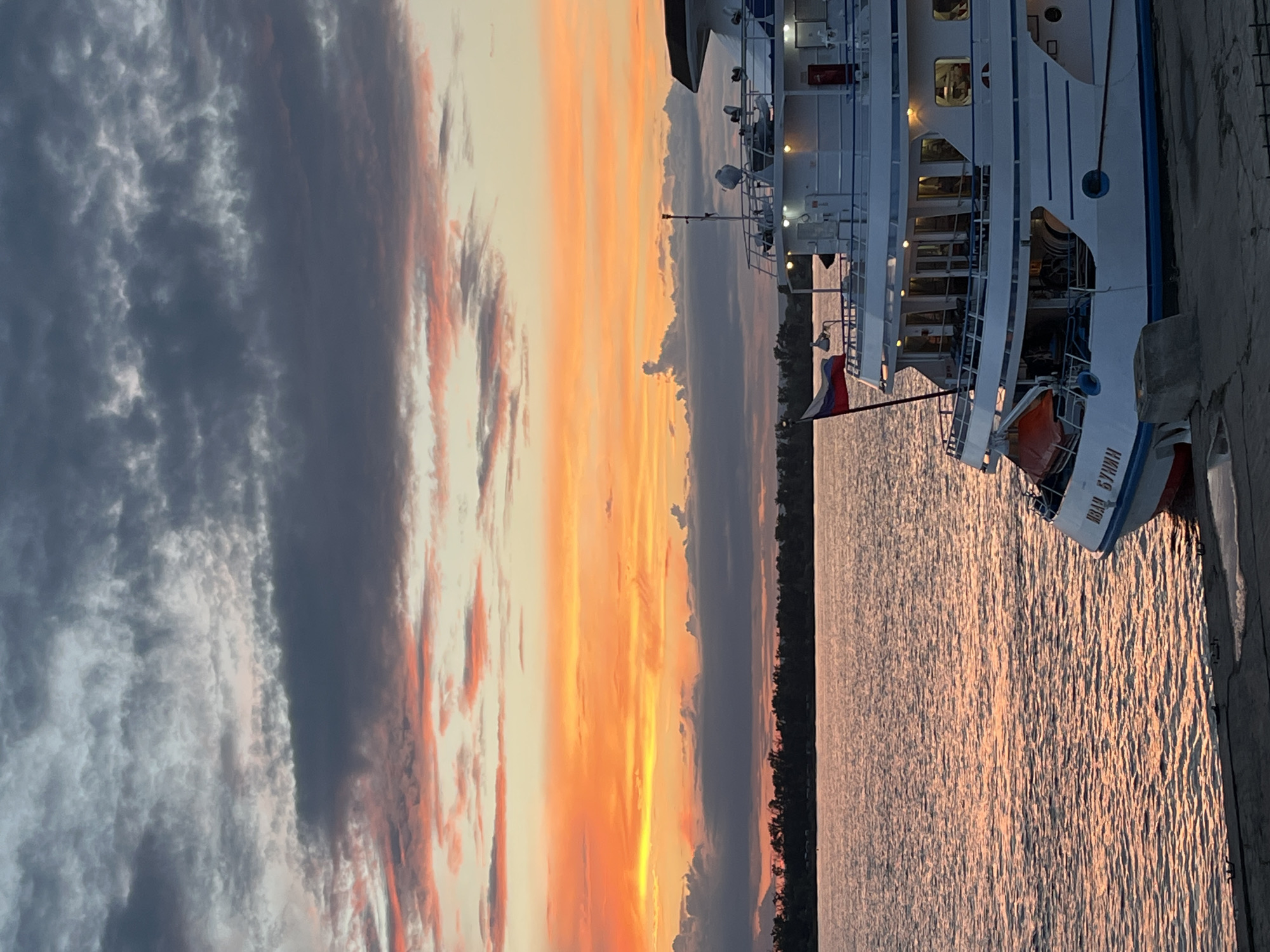 A white passenger ship with a Russian flag is docked on the shore of a river at sunset. The sky is filled with vibrant orange, pink, and gray clouds, reflecting on the calm water. The ship has multiple decks with lit windows, suggesting it is occupied. The name "ИВАН БУНИН" (Ivan Bunin) is visible on the hull. A treeline of dark trees forms the distant shore. The scene appears to be located along the Volga River, potentially near Novokuybyshevsk, Russia, given the ship's name, which is a common name for vessels operating in that region.