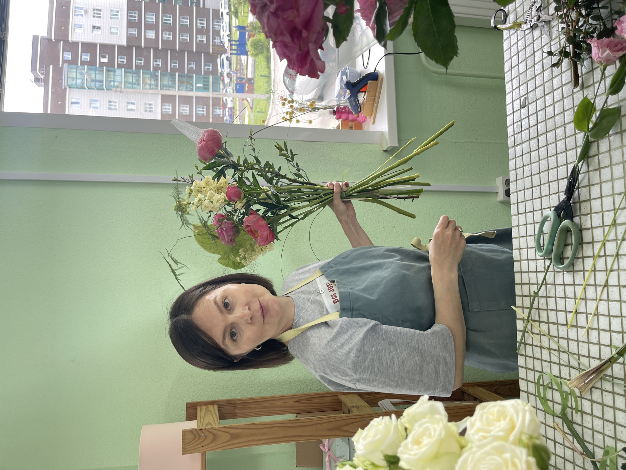 A woman stands in a room with light green walls, holding a bouquet of pink and yellow flowers. She is wearing a grey t-shirt and a blue apron over it, and her short brown hair frames her face. She is looking towards the camera with a slight smile. In the background, a window reveals a cityscape with tall apartment buildings and a playground. On a table in front of her, a checkered white surface is strewn with various floral accessories, including scissors, a hot glue gun, and loose flowers. The scene appears to be a flower shop or a workspace dedicated to floral arrangement.