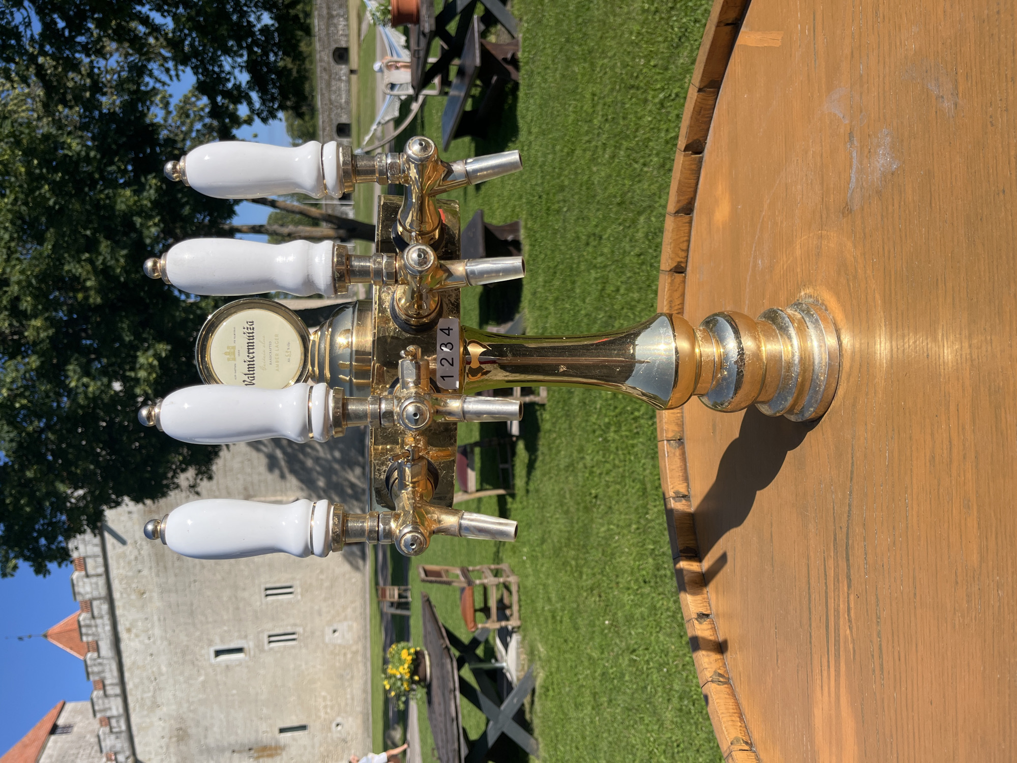 The image shows a golden beer tap with four white handles, positioned on a wooden surface, likely a barrel top. In the background, a stone castle wall with red-tiled roofs and crenellations is visible, bathed in sunlight under a clear blue sky. Trees with lush green foliage are also present. The castle is identifiable as Kuressaare Castle, located in the city of Kuressaare, Estonia. The setting appears to be an outdoor area, possibly a courtyard or garden adjacent to the castle, with grass, tables, chairs, and a hammock visible, suggesting a relaxed, possibly tourist-oriented environment. One person, a woman in a white dress, is seen walking in the distance on the left side of the frame. The focus is on the beer tap, rendered in sharp detail, contrasting with the softer focus of the background.