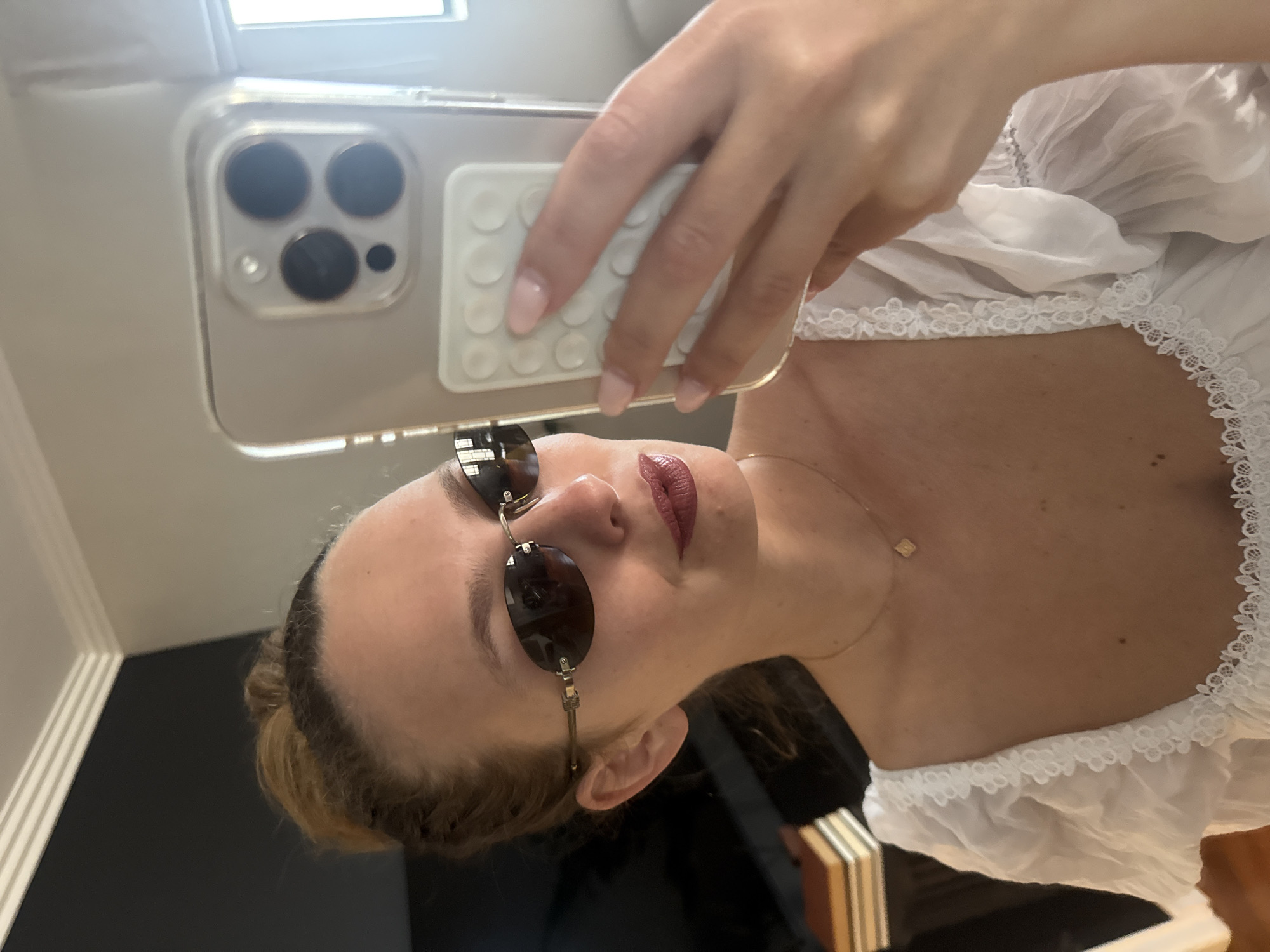 The image depicts a selfie taken with a smartphone. The subject, a woman, is wearing oval sunglasses and a white top with lace trim. She has a gold necklace with a small square pendant. Her lipstick appears to be a dark red or burgundy shade. The phone is held in her hand, with a phone case featuring a circular grip pad. The background is partially visible, showing a black wall, a wooden frame, and part of a white window frame. There is no evidence to suggest the location is Cape Town, South Africa.