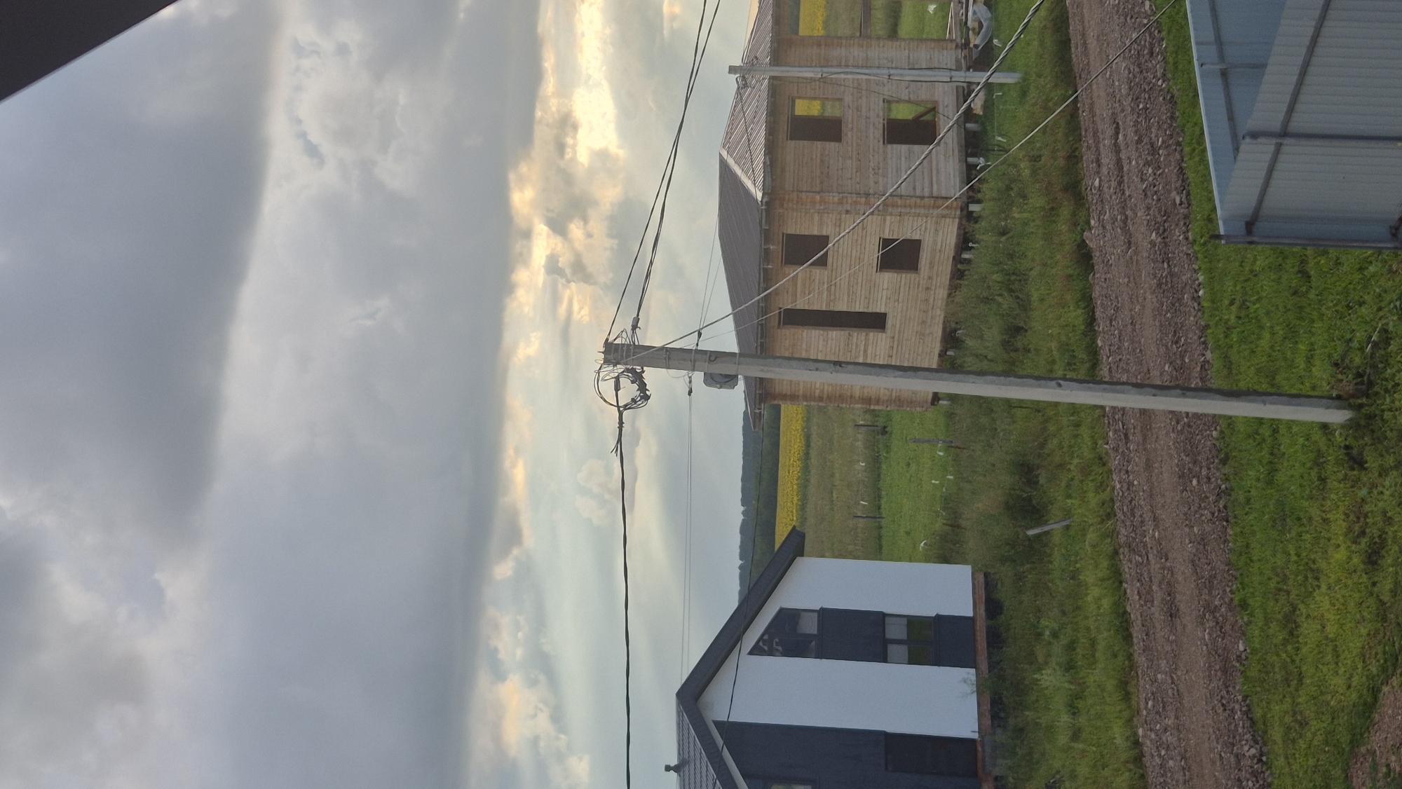 The image depicts an exterior view of houses under construction in Pomryaskino, Russia, under a cloudy sky. In the foreground, a dirt road runs through a grassy area, with a light gray fence visible at the bottom right. A concrete utility pole stands prominently in the center, supporting several electrical wires that stretch across the frame. To the left, a house with a white and dark gray exterior is partially visible. To the right, a two-story wooden house, seemingly under construction, is featured. In the background, a field of yellow flowers, possibly sunflowers, stretches towards a line of trees. The overall scene suggests a rural or semi-rural setting with ongoing development.