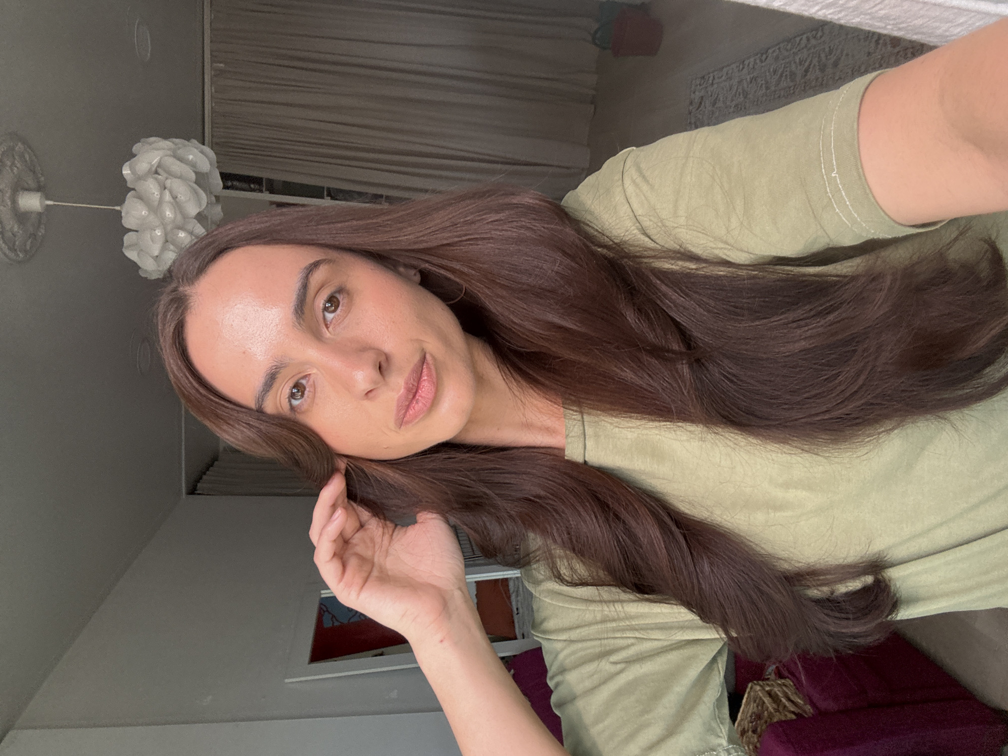 A woman with long, brown hair and an olive-green t-shirt is pictured in a close-up selfie. The background shows a room with a white ceiling light fixture, beige curtains, and a partial view of a room with a gray wall. Details that can indicate the location of the city of Krasnoyarsk are not provided by the image.