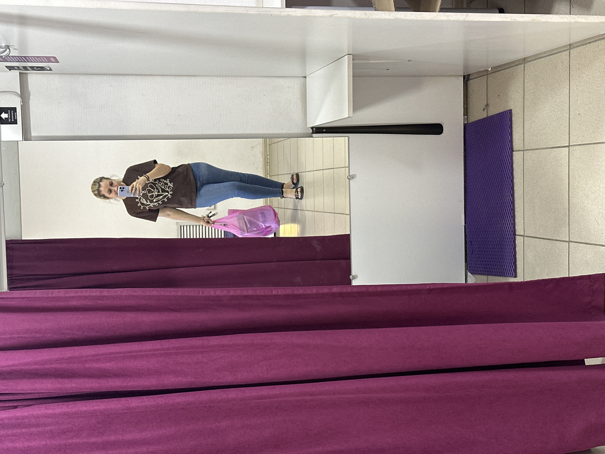 The image shows a woman standing in a fitting room in a store in Novoaltaysk, Russia. She is holding a pink plastic bag and a cell phone. She is wearing a brown t-shirt and blue jeans. The fitting room has a purple curtain on the left side, and a mirror on the right side. There is a shelf inside the fitting room, and a purple mat on the floor. The floor is tiled.