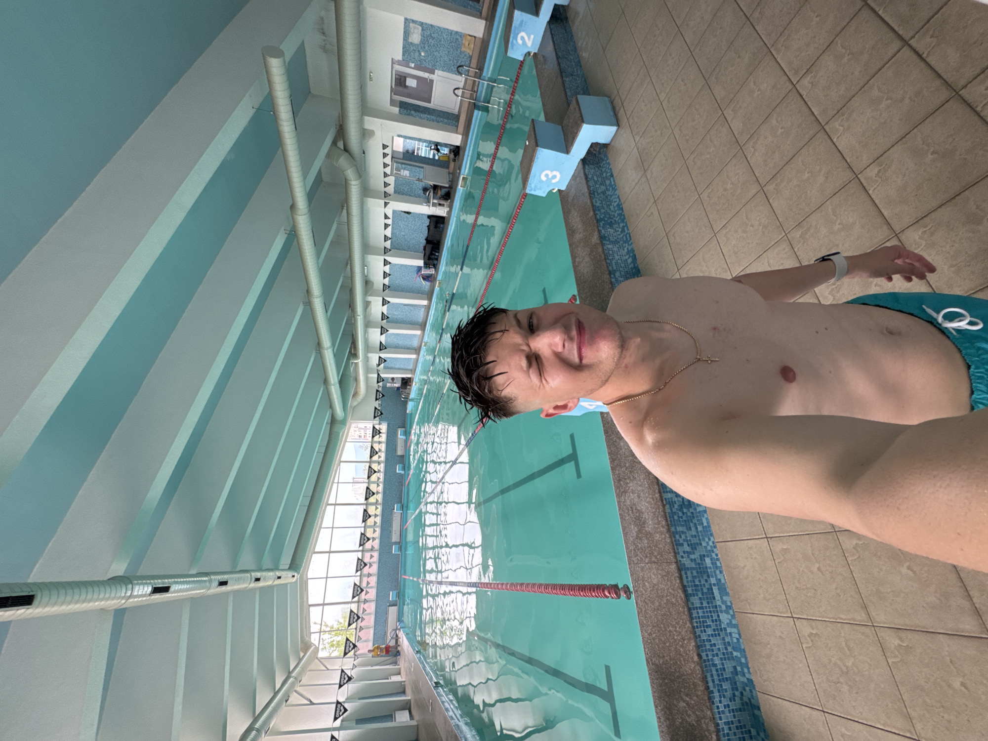 A selfie captures a young man, shirtless and wearing turquoise swim shorts, standing beside an indoor swimming pool, likely in Plovdiv, Bulgaria. His hair appears wet, suggesting he has recently been swimming. He displays a slight, squinted smile, possibly due to bright lighting or the act of taking the photo. A gold chain with a cross pendant is visible, along with a white smartwatch on his left wrist.
The scene is an indoor swimming complex, well-lit by natural light streaming through large windows along one side of the pool, revealing glimpses of trees and buildings outside. The long pool features clear turquoise water with several lanes marked by red and white ropes. Two starting blocks, clearly numbered "2" and "3", are visible at the near end of the pool. The pool deck is tiled in a light brown pattern.
Architecturally, the facility features walls painted with alternating light blue and white horizontal stripes and multiple silver ventilation ducts running along the ceiling and upper walls. Black and white triangular flags, some bearing the "ARENA" logo, are strung across the pool area. A red lifeguard chair can be seen in the distance next to the windows. The environment suggests a functional and modern swimming facility, likely used for training or recreation during the daytime.
