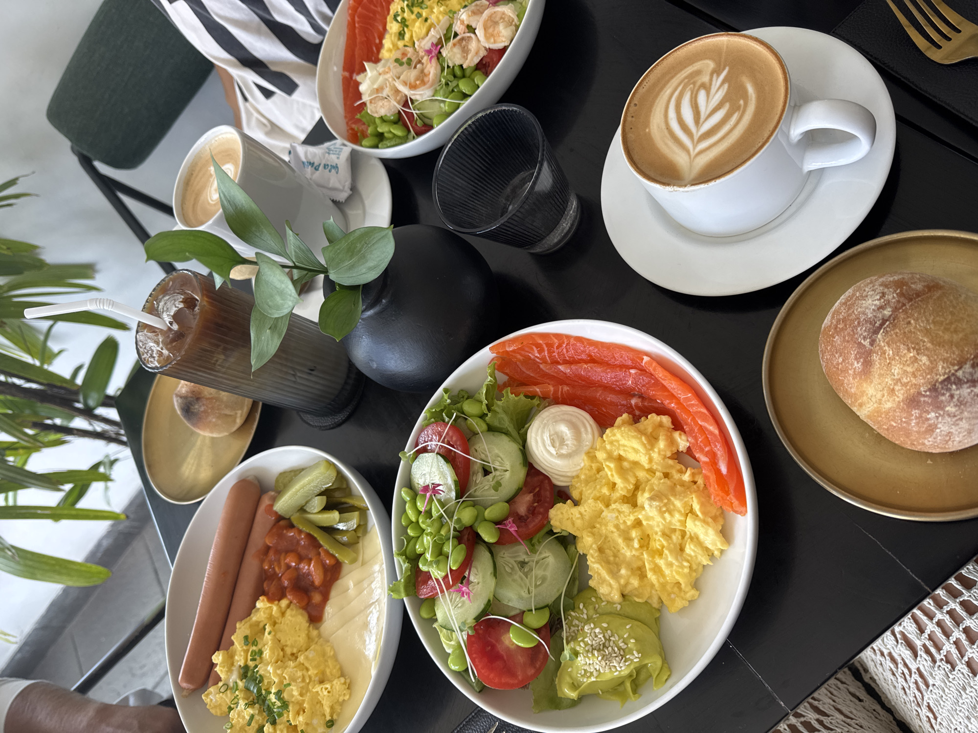 A vibrant overhead shot captures a rich breakfast or brunch spread laid out on a sleek, dark table, likely black marble. The scene suggests a relaxed meal for at least two people, with diverse dishes and beverages arranged artfully.
In the foreground, a white oval plate holds two hot dogs, a dollop of baked beans, several pickled gherkins, and slices of what appears to be cheese, alongside a generous portion of fluffy scrambled eggs garnished with chives. Adjacent to this, another white oval plate features neatly arranged slices of smoked salmon, a fresh green salad with lettuce, cucumber, and tomato, green edamame beans, a swirl of white sauce (possibly mayonnaise or a similar dressing), creamy mashed avocado topped with sesame seeds, and more scrambled eggs.
Towards the upper left, a white bowl contains another salad with salmon, several cooked shrimp, edamame, and various greens, indicating a separate, lighter meal option or an additional sharing dish.
Beverages include two white ceramic cups of coffee, both featuring intricate latte art, placed on matching saucers. One cup is positioned in the upper right, and another is partially visible in the upper left. A tall glass with ice and a dark beverage, possibly iced coffee or tea, is seen with a white straw, accompanied by a small black vase holding green foliage. An empty, fluted dark glass also sits on the table.
A golden-colored round plate holds a crusty bread roll. The table is adorned with a small black vase with green leaves, adding a touch of natural elegance. Portions of a person's arm and clothing, including a white and black striped top and a lacy white fabric, are visible at the edges of the frame. A small white packet with partially visible text "Gula Pali" (likely sugar) is also on the table. The lighting is bright and natural, indicating daytime, possibly in a cafe or restaurant given the professional presentation of the food.