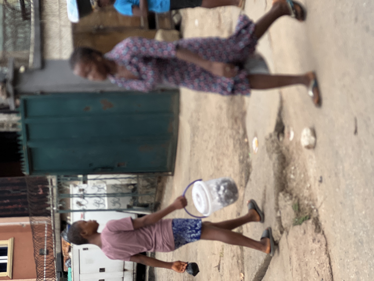 This image captures a street scene in Nkpor, Nigeria, during daylight hours, featuring three individuals walking. The overall composition shows a bustling, lived-in environment.
In the foreground, a young person, likely a girl, walks towards the left side of the frame (if oriented upright). She is wearing a light pink t-shirt, blue patterned shorts, and dark flip-flops. She carries a white bucket with a translucent lid, possibly containing goods for sale or personal items, in her right hand. Her gaze is directed forward.
Further into the mid-ground, another young person, also appearing to be a girl, walks in the same direction. She is dressed in a patterned, long-sleeved dress with shades of purple and pink, and light-colored flip-flops. Her figure is slightly blurred, suggesting motion. In the background, a third person is partially visible, wearing a blue t-shirt and dark bottoms.
The setting is an outdoor street or pathway, paved with concrete or asphalt, showing some signs of wear with cracks and scattered debris. Buildings line the street, including one visible on the left with a white window frame and a section of barbed wire fencing. A large, white, generator-like appliance is situated behind the foreground person. A prominent dark green metal gate, likely an entrance to a property, stands closed in the middle ground. The scene depicts everyday activity under clear skies. No visible text is discernible in the image.