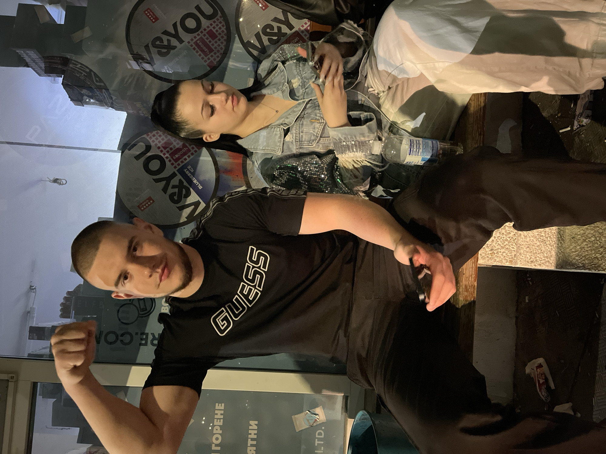 The image depicts a man and a woman sitting near a storefront with advertising for V&YOU, likely in Burgas, Bulgaria, given the Cyrillic text ("ГОРЕНЕ," "ЯТНИ," "LTD.") visible on the storefront. The man is wearing a black "GUESS" t-shirt and dark pants, flexing his bicep for the camera. The woman is reclining behind him, wearing a denim jacket with sequin detailing and light-colored pants. She is holding a smartphone. A bottle of water is visible near her. The storefront window reflects street elements, including buildings.