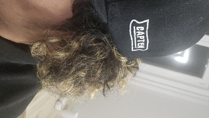 A person with shoulder-length, wavy, light brown hair with blonde highlights wears a dark-colored baseball cap with the word "CAPTEN" embroidered in white. The person is tilted to the side, revealing part of their neck and shoulder, which are covered by a dark-colored garment. The background consists of a white wall and a section of light-colored flooring. A white cylindrical object is visible near the bottom left corner.