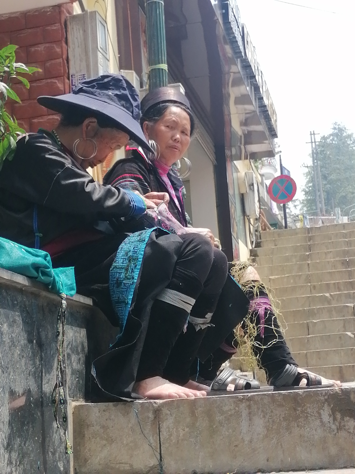 Two women sit on a stone ledge next to a set of stairs in Sa Pa Ward, Vietnam. The woman on the left wears a wide-brimmed dark hat, a black top with blue trim, black pants, and large silver hoop earrings; she is barefoot. Next to her sits a woman wearing a traditional headpiece, a black and pink top, black pants, large silver hoop earrings, and sandals. A blue bag hangs from the ledge next to the first woman. The stairs lead up to a building with a “no parking” sign.