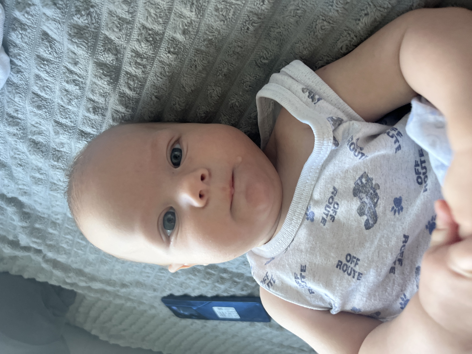 A close-up, eye-level shot shows a baby lying on its back on a grey, textured blanket. The baby, wearing a white onesie with a repeating "OFF ROUTE" pattern and small paw prints, is positioned with its head turned towards the right of the frame. The baby's left eye is clearly visible, a striking blue, while its right eye is partially obscured. A single tear or droplet of saliva is visible on the baby's chin. The background shows a portion of a dark blue smartphone lying on the blanket. The lighting appears natural, coming from the left, casting soft shadows. The scene is indoor, likely a bedroom or living area.
