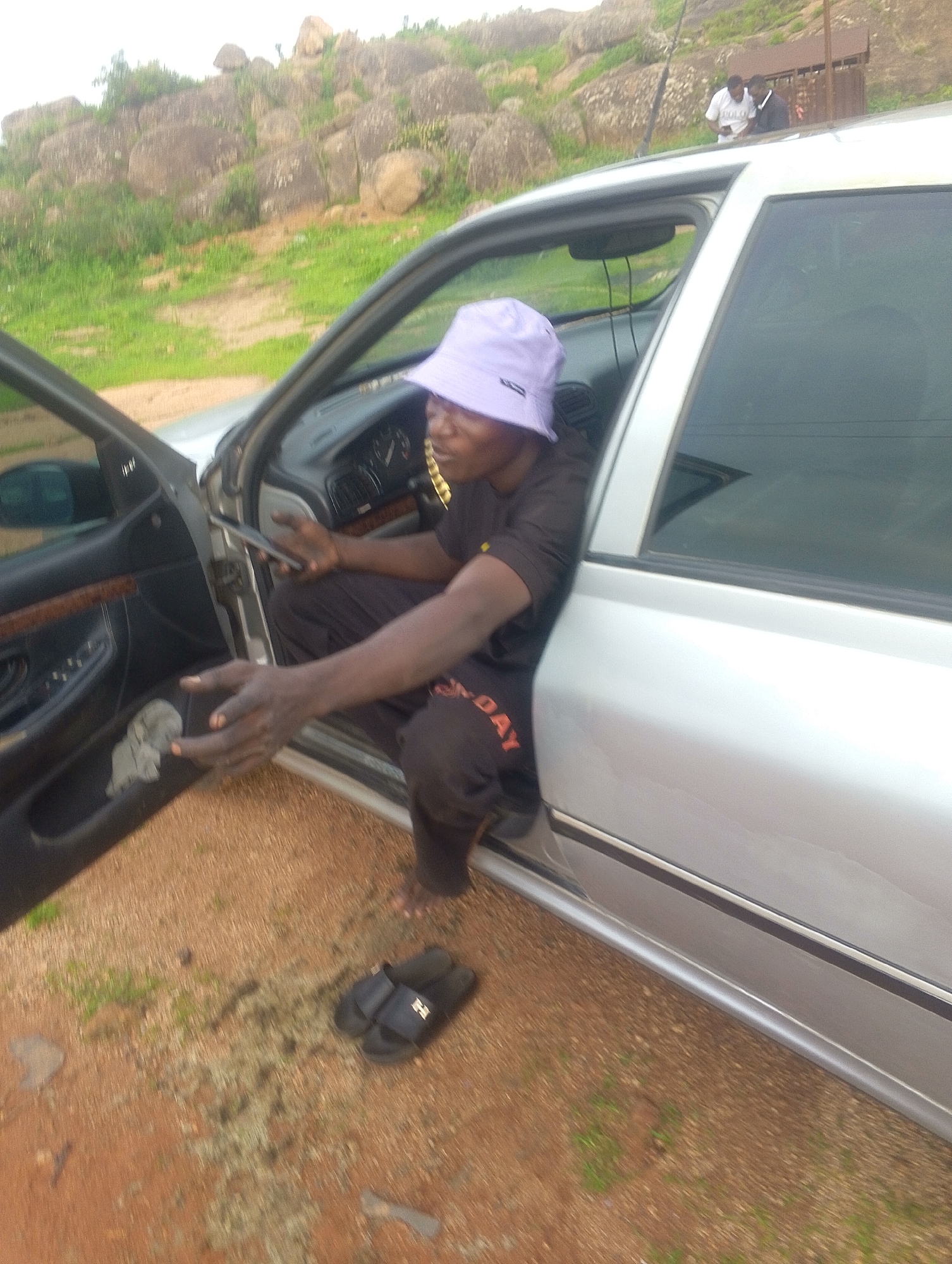 In Jos, Nigeria, a man wearing a light purple bucket hat and a dark t-shirt with orange writing on his pants sits in the driver's seat of a silver sedan, with his upper body leaning out of the open driver's door. He is holding a mobile phone in his left hand. His right arm is extended forward. A pair of black flip-flops rests on the reddish-brown, gravelly ground outside the car door. In the background, a rocky, vegetated hillside rises, with two other individuals standing on a brown structure in the distance, on top of the car.