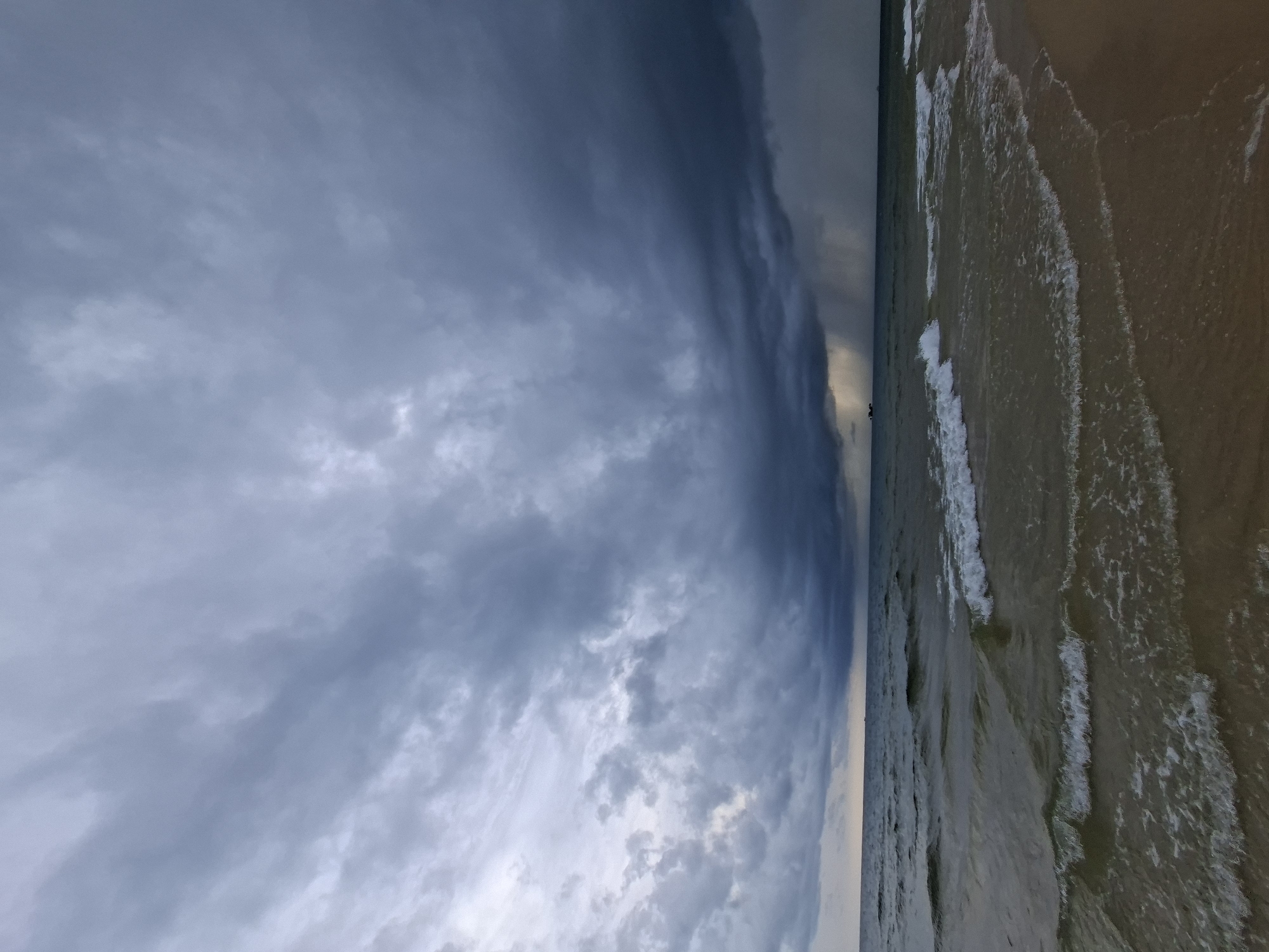 A beach scene is captured under a dramatic, stormy sky. Dark, heavy clouds dominate the upper portion of the image, with lighter patches of sky visible on the horizon. Towards the right side of the frame, a distinct rain shaft is visible falling from the clouds onto the sea. The water is choppy, with whitecaps on the waves breaking near the shore. The beach itself appears to be sandy and wet. In the distance, on the horizon, a few small, indistinct shapes may be boats or structures. The overall mood is one of impending or ongoing bad weather.