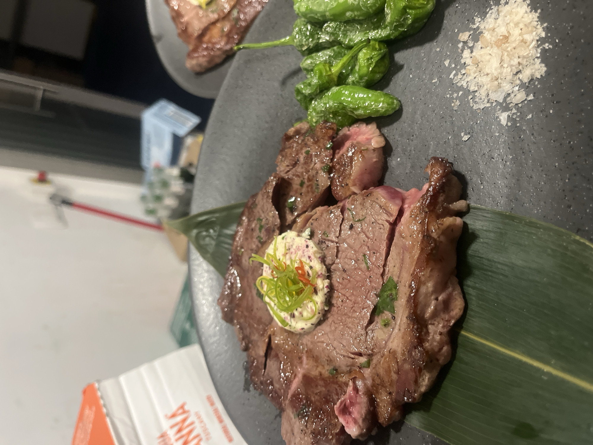 The image shows a close-up shot of a plate of food, likely taken indoors. The main subject is a piece of grilled meat, possibly steak, served on a green leaf. Atop the meat is a dollop of what appears to be butter or cream cheese, garnished with finely chopped green and red herbs. To the side of the meat are several small, green peppers, likely shishito peppers, and a small pile of coarse salt crystals. The plate is dark grey, and a portion of another plate with a similar dish is visible in the background. Based on the presence of a package labelled "ANNA" - likely a brand of food product - and the style of the dish, it's probable the photo was taken in a restaurant. Without further information or context embedded in the image, it is not possible to determine the exact location in Sofia, Bulgaria.