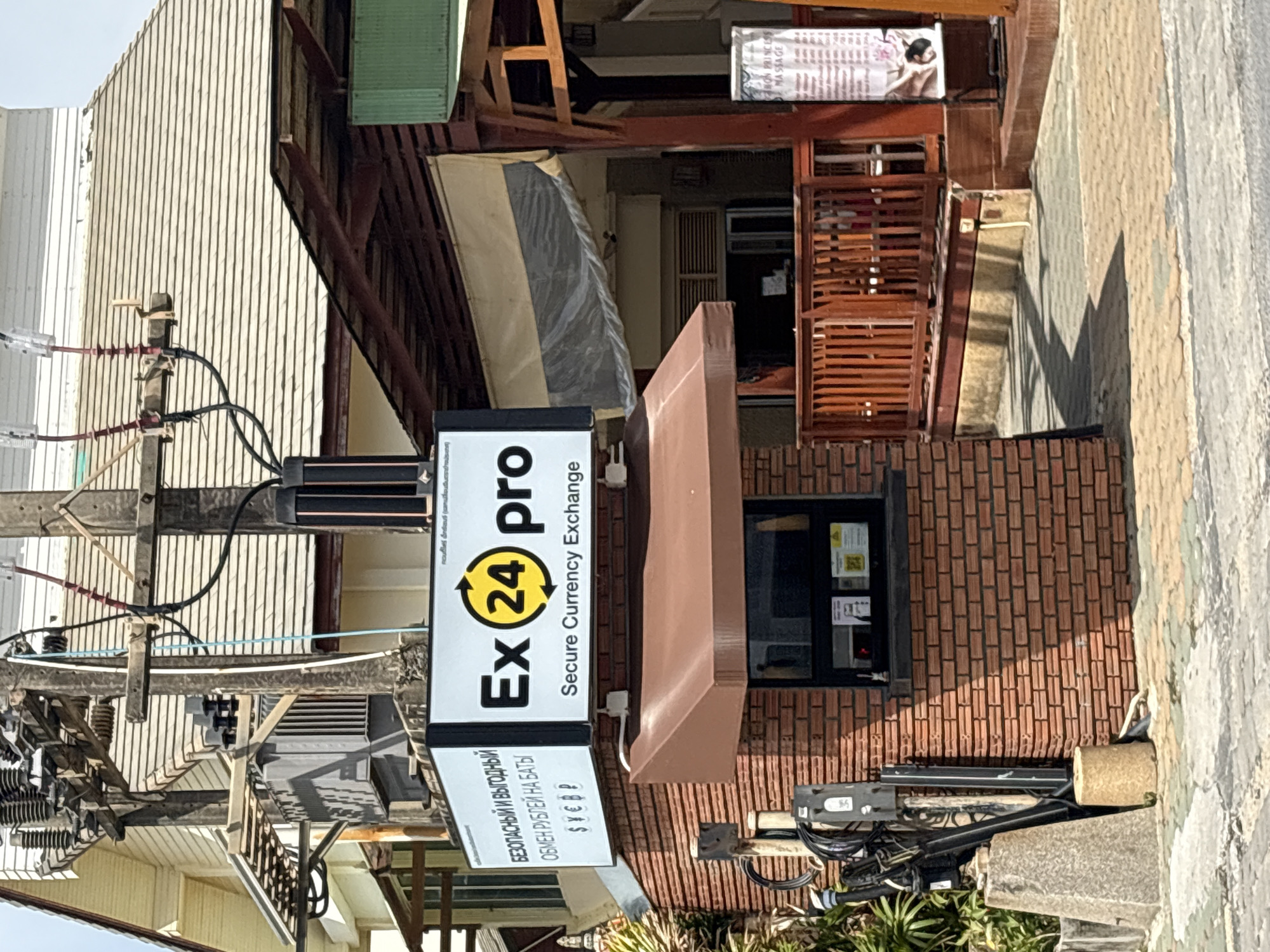 A currency exchange sign is mounted on a pole in front of a brick building. The sign reads "Ex24pro Secure Currency Exchange" in English and Russian. The building has a small window where transactions may occur, and a brown awning extends over the window. To the right of the building, a wooden fence surrounds an area with tables and chairs, suggesting an outdoor seating area for a business. A sign advertising "THAI PRINCESS MASSAGE" is visible on the right side of the image. Electrical wires and transformers are present on the pole, indicating the area is served by public utilities. The ground is paved with interlocking bricks and concrete. Lush green foliage is visible to the left of the building. The scene appears to be in a commercial area, likely in Karon, Thailand, given the presence of Thai script on some signs and the overall ambiance. There are no people visible in the image.